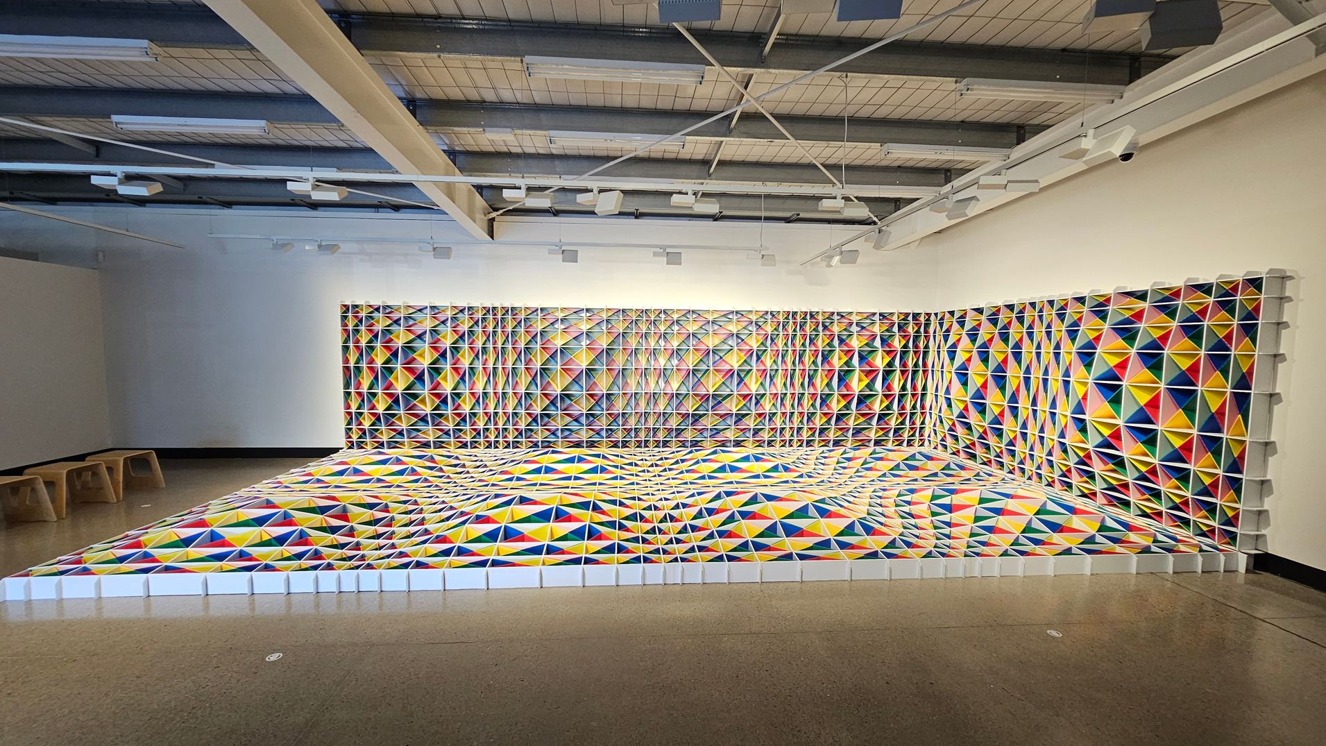 A room with a geometric art installation covering the floor and two walls. The artwork consists of colourful triangular patterns in red, blue, yellow, and white, creating a three-dimensional effect. The ceiling has exposed beams and lighting fixtures. On the left, there are wooden benches along the wall. The floor is polished concrete, and the walls are painted white, enhancing the vibrant colours of the installation.