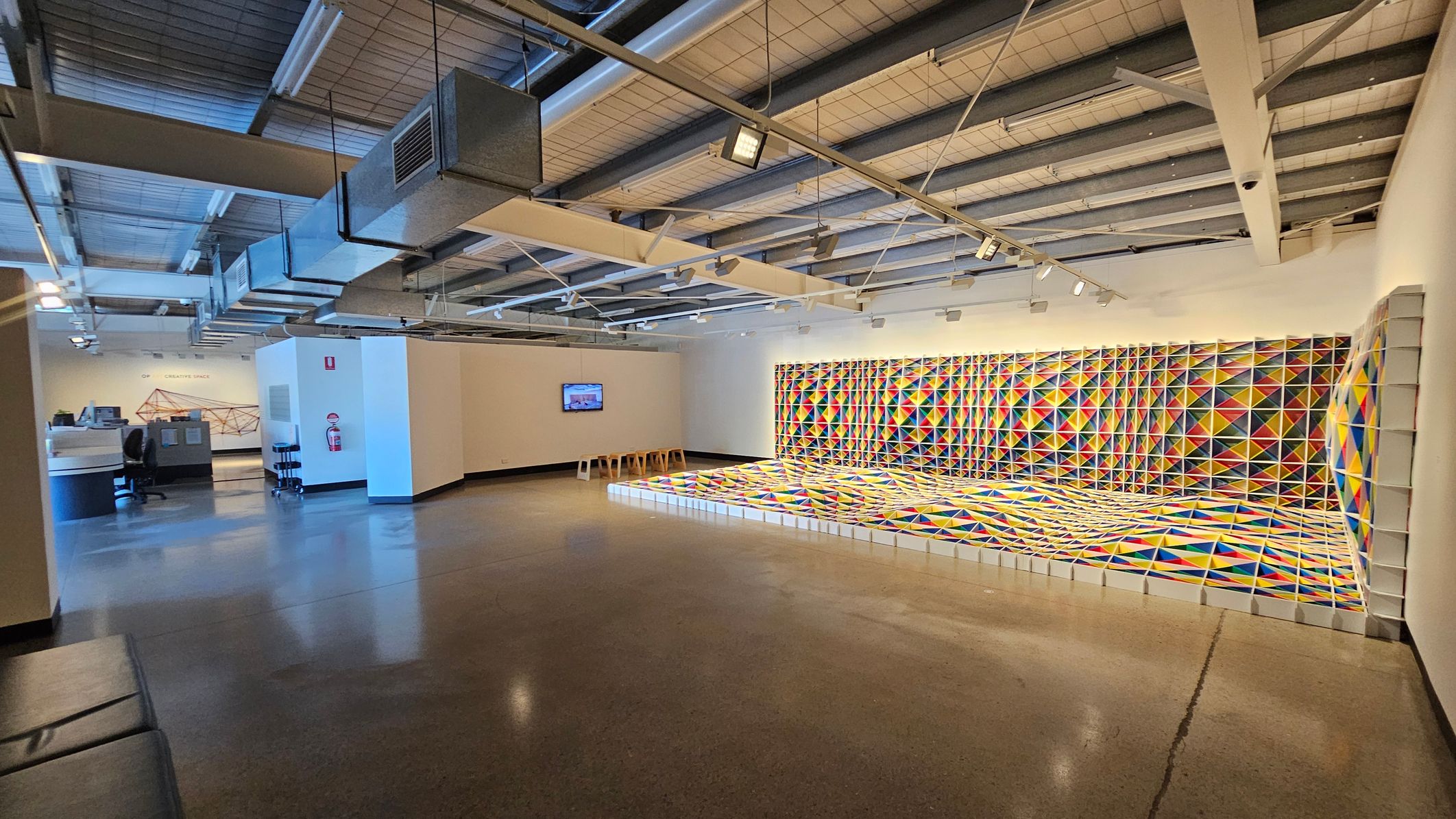 A spacious room with a polished concrete floor and an industrial-style ceiling featuring exposed beams and ductwork. On the right, a vibrant, geometric art installation with colourful triangular patterns covers the wall and extends onto the floor. In the middle, a white wall with a mounted screen and several stools. On the left, a desk with office chairs and a wall with a geometric design. The lighting is bright, highlighting the artwork.