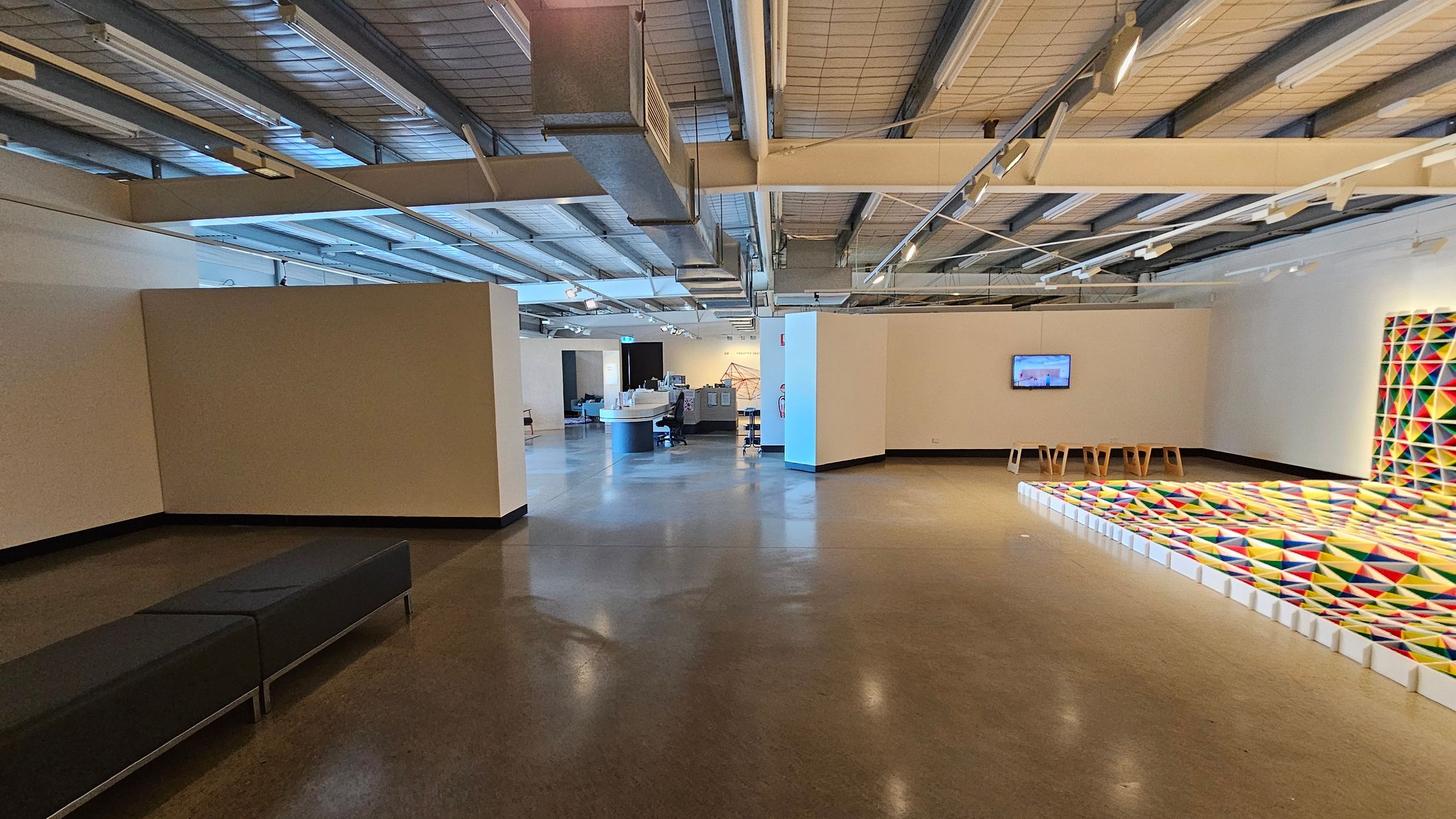 A spacious interior with a polished concrete floor and an industrial-style ceiling featuring exposed beams and ductwork. On the left, there is a long, dark bench against a plain wall. In the centre, a reception desk with a person seated behind it is visible. On the right, a colourful geometric art installation is displayed on the floor and wall, with a small screen mounted on the wall above a few wooden stools. The area is well-lit with overhead lights.