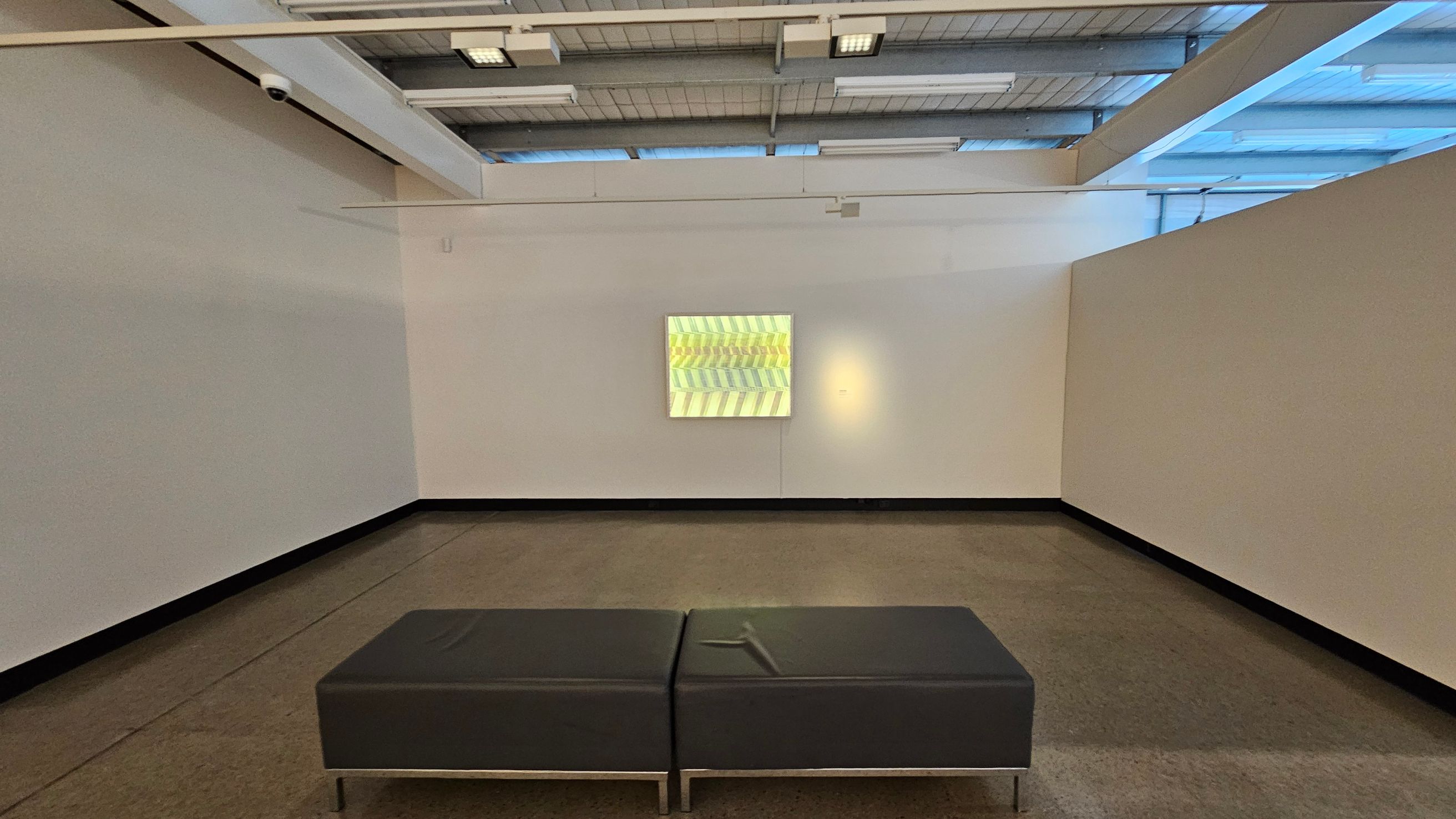 A minimalist gallery space with a grey bench in the foreground on a polished concrete floor. The walls are plain and light-coloured, with a single piece of abstract art featuring a pattern of yellow and green chevrons in the centre. The ceiling is industrial with exposed beams and fluorescent lighting. A security camera is mounted on the left wall. The space is well-lit, with natural light coming from windows near the ceiling.