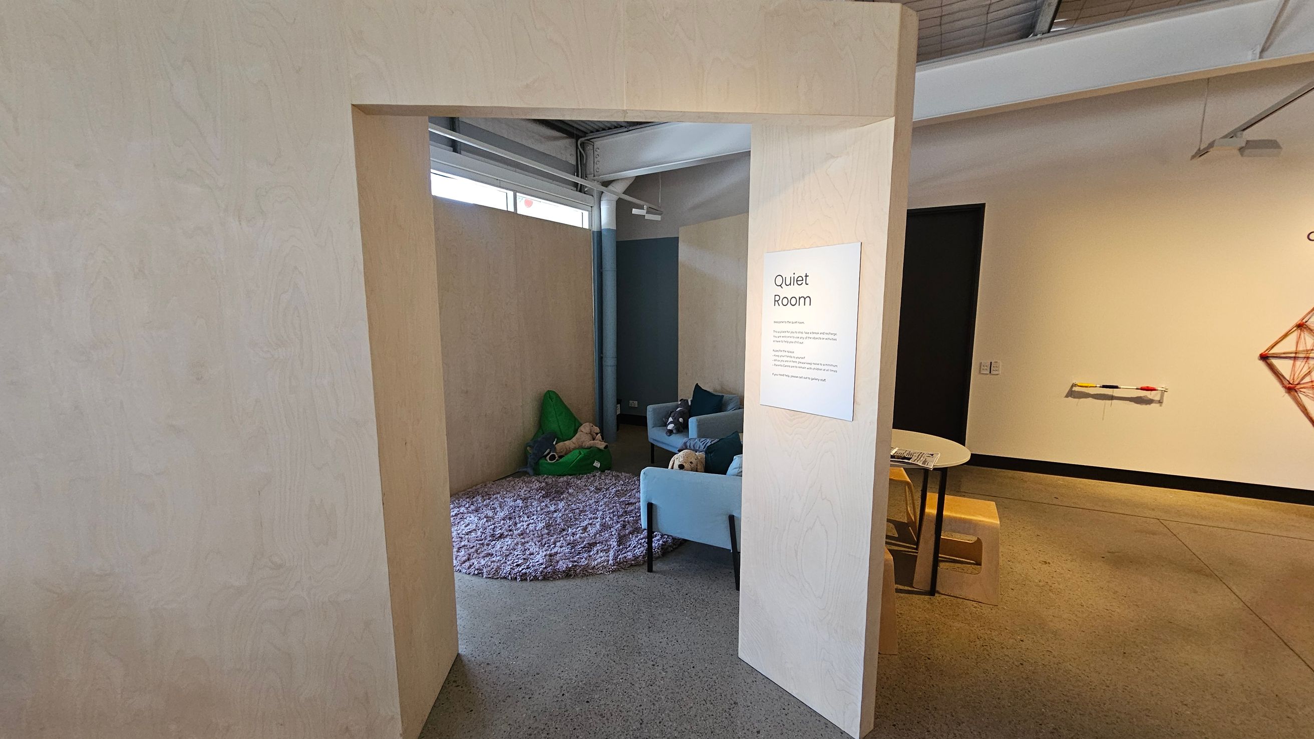 A room with a sign labelled "Quiet Room" on the right wall. Inside, there is a light blue sofa with cushions and a plush toy. A green bean bag and a round, textured rug are on the floor. The walls are light-coloured, and the ceiling has exposed beams. In the foreground, a small table with chairs is visible, and a geometric wall decoration is on the right. The floor is polished concrete.