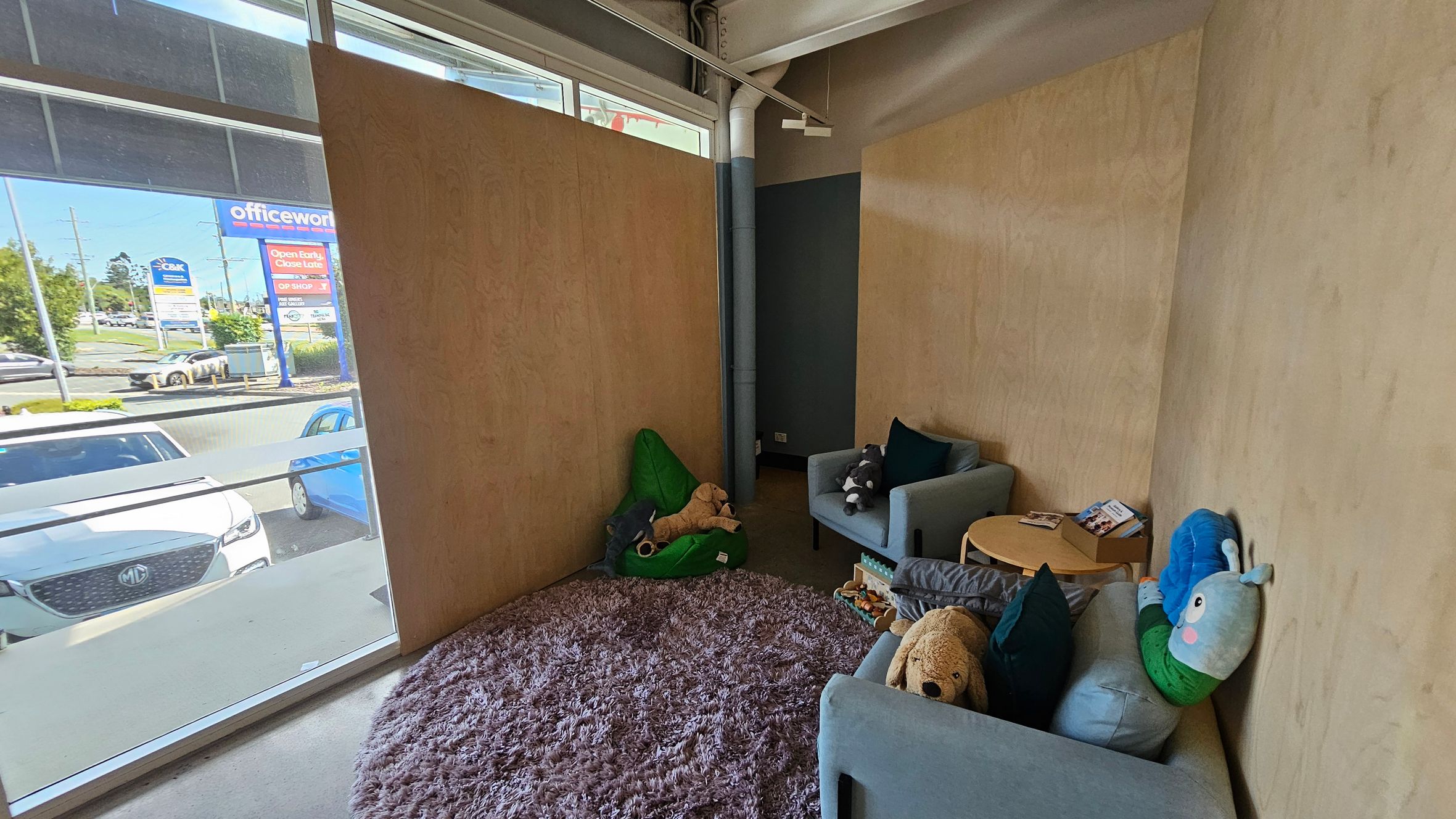 A cosy room with light wooden walls and a large window on the left, showing a view of parked cars and a street with signs. Inside, there are two grey armchairs with plush toys, including a dog and a caterpillar. A small round wooden table holds a few books. A green bean bag with a stuffed animal is on the floor, which is covered by a fluffy purple rug. The ceiling is industrial with exposed pipes.