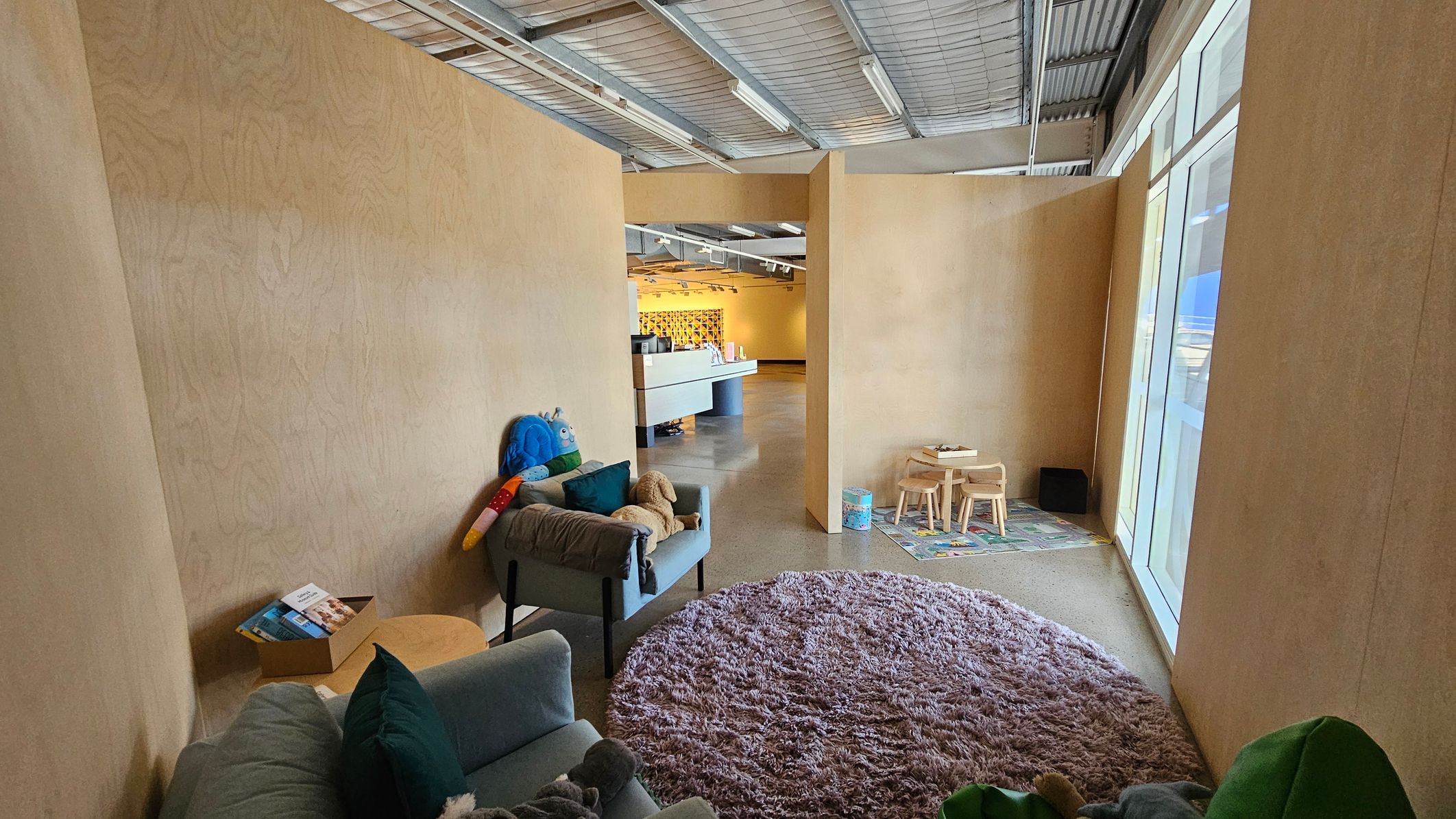 A room with light wooden walls and a concrete floor. On the left, there's a small sofa with green cushions and a plush toy, next to a cardboard box with brochures. In the centre, a purple shaggy rug covers the floor. On the right, a children's play area with a small table, chairs, and a colourful mat. Large windows line the right wall, allowing natural light in. The ceiling is industrial with exposed metal beams and fluorescent lights. In the background, a reception desk is visible.