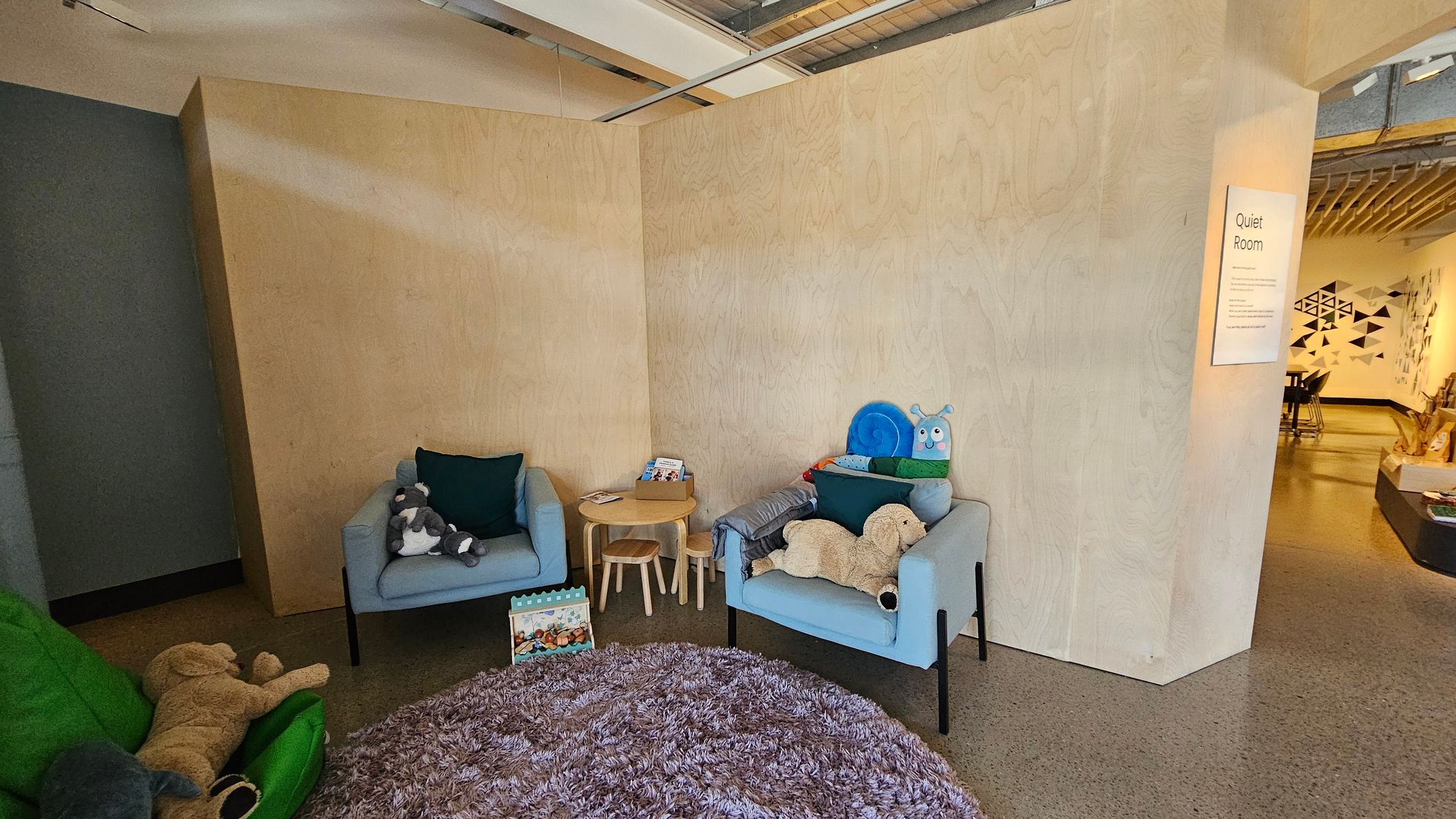 A cosy room with light wooden walls. On the left, a green chair with a large teddy bear and a grey stuffed animal. In the centre, two light blue armchairs with dark green cushions, one holding a stuffed elephant and the other a plush caterpillar and dog. A small round wooden table with children's books and stools is between the chairs. A fluffy purple rug covers the floor. On the right, a sign reads "Quiet Room" near an open entrance to another area with geometric wall art.