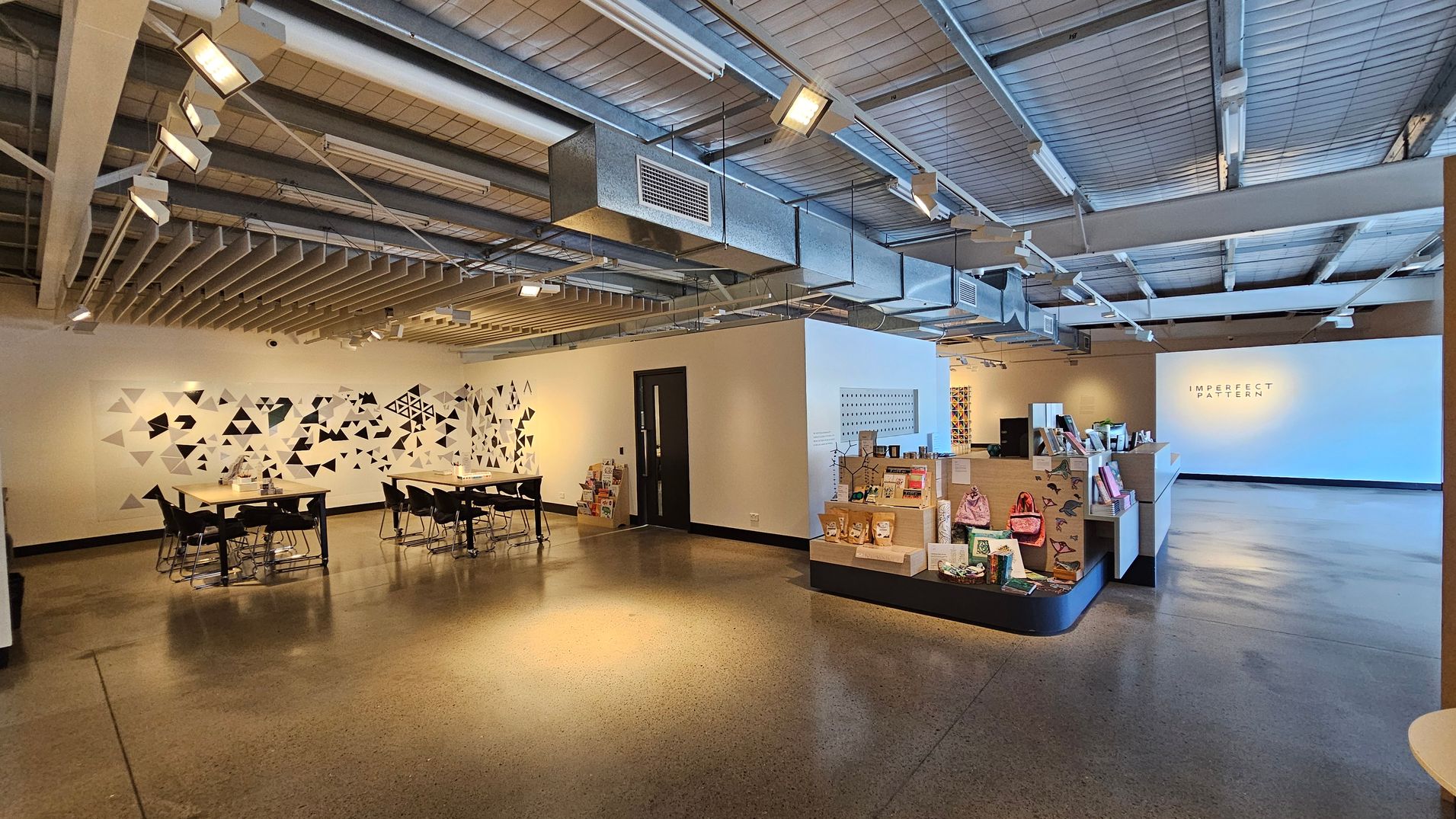 A spacious interior with a polished concrete floor and an industrial-style ceiling featuring exposed ducts and lighting. On the left, there are tables and chairs near a wall adorned with a geometric black and white triangle pattern. In the centre, a small display area showcases various items, including books and bags. On the right, a large white wall displays the text "IMPERFECT PATTERN." The overall atmosphere is modern and open.