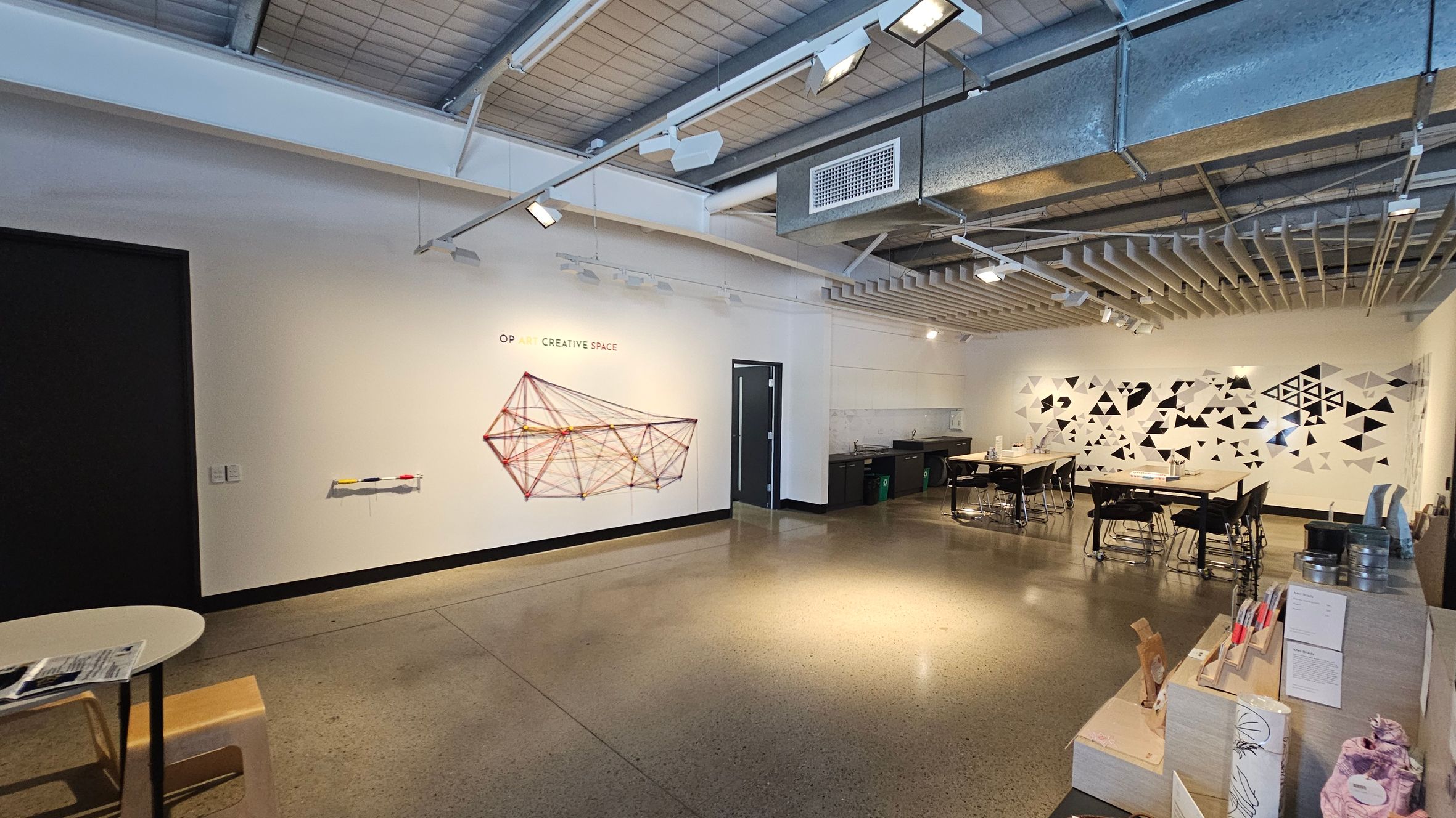 A spacious room with a polished concrete floor and an industrial-style ceiling with exposed ducts and lighting. On the left wall, there's a geometric string art installation labelled "OP ART CREATIVE SPACE." To the right, a wall features a pattern of black and white triangles. Several tables with chairs are arranged in the middle, and a counter with various items is on the far right. A small table and stool are in the foreground on the left.