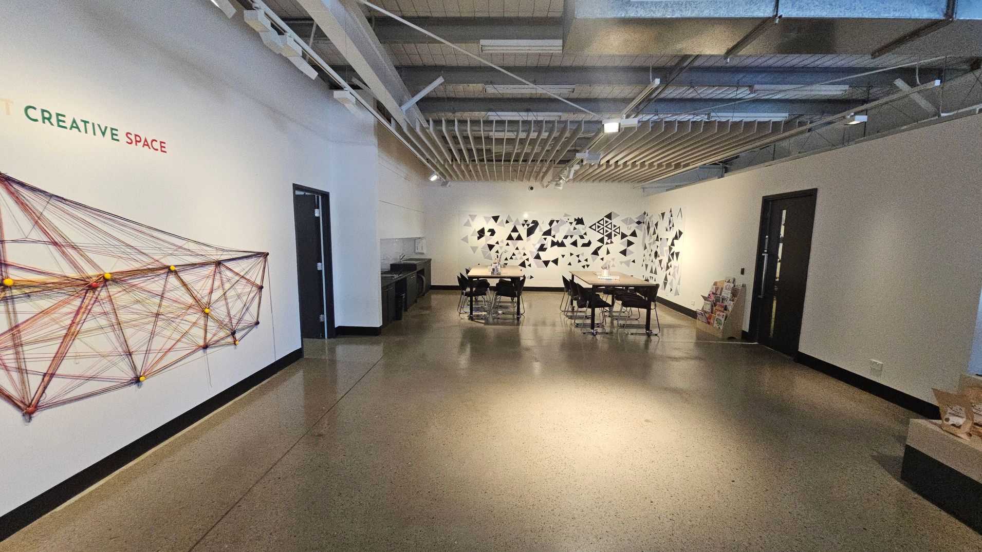 A room with a polished concrete floor and an exposed ceiling with beams and ducts. On the left wall, there's a geometric string art installation with the words "CREATIVE SPACE" above it. In the centre, two tables with chairs are arranged, and the back wall features a pattern of black and white triangles. On the right, there are two black doors and a stack of books or magazines on a low shelf.