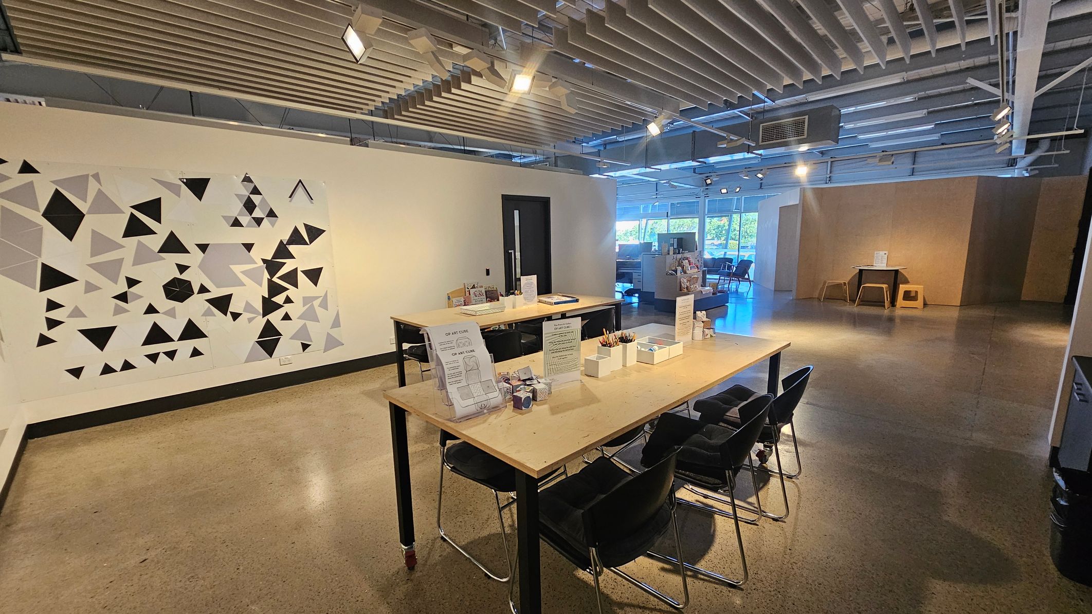 A modern interior space with a polished concrete floor and a ceiling featuring exposed beams and spotlights. On the left, a wall displays a geometric pattern of black and white triangles. In the centre, two wooden tables with black chairs are arranged, holding various papers, pens, and informational displays. The background shows a large window with natural light streaming in, and a small table with stools is visible against a plain wall.