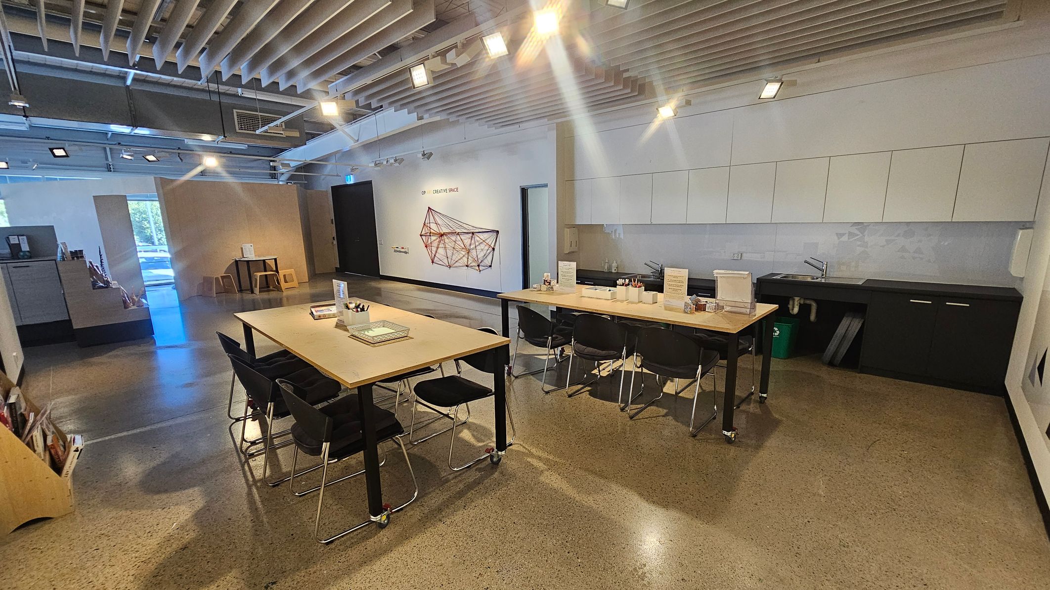 A modern workspace with a polished concrete floor and a ceiling featuring exposed beams and lights. On the right, there are white cabinets above a black countertop with a sink. Two long tables with black chairs are in the centre, equipped with stationery and informational displays. On the left, a small table and stools are near a window, and a shelf holds books or magazines. A geometric wall art piece is visible in the background.