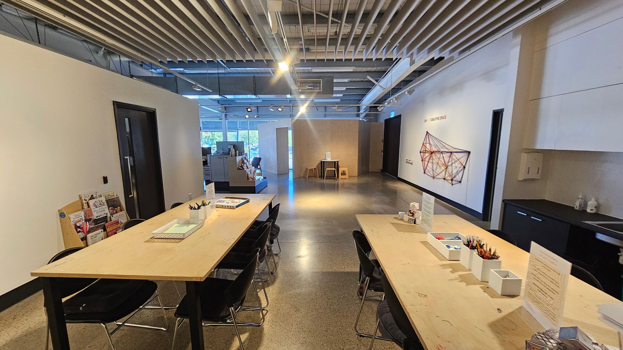 A spacious room with a polished concrete floor and a ceiling featuring exposed beams and lights. On the left, there are wooden tables with black chairs, books, and stationery. The right side has a wall with a geometric string art piece and a counter with supplies. In the background, a small table with stools is near large windows, allowing natural light to enter. The room has a modern, open design with a focus on creativity and learning.