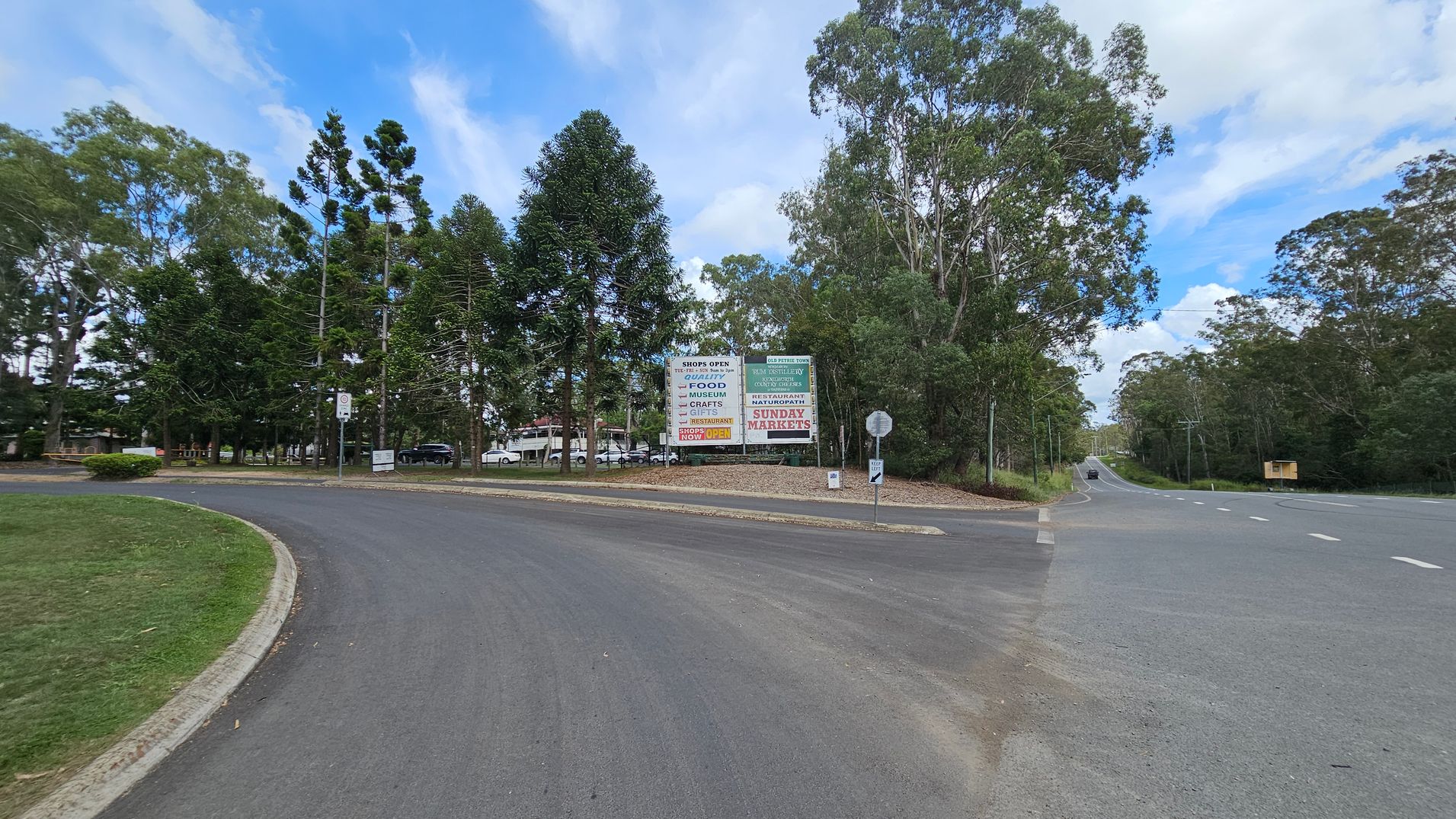 A road curves to the left with a grassy verge on the left side. In the centre, there are tall trees and a large sign advertising shops, food, and Sunday markets. To the right, a straight road extends into the distance, flanked by more trees. A small sign and a bin are near the road junction. The sky is partly cloudy.