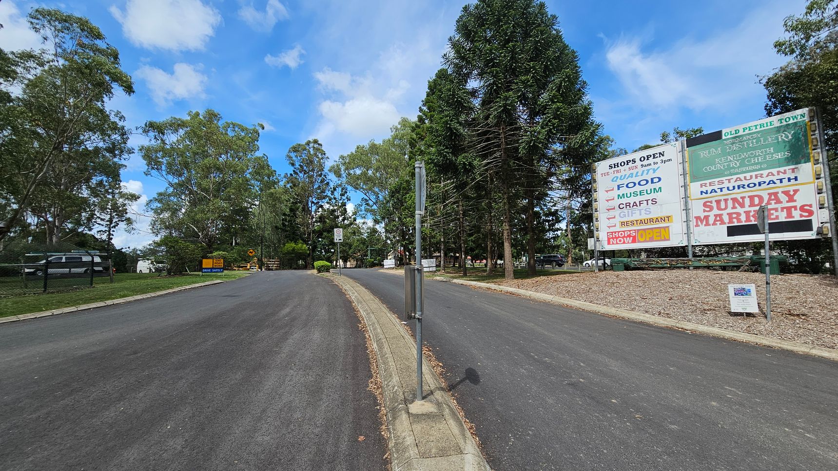 A road with a central dividing strip leads into a tree-lined area. On the right, a large sign advertises shops, a museum, crafts, gifts, a restaurant, and Sunday markets. The left side features a grassy area with a fence and parked cars. Tall trees are visible on both sides, and a smaller sign on the left reads "Drive Safely." The road surface is dark and smooth, with a kerb separating the lanes.