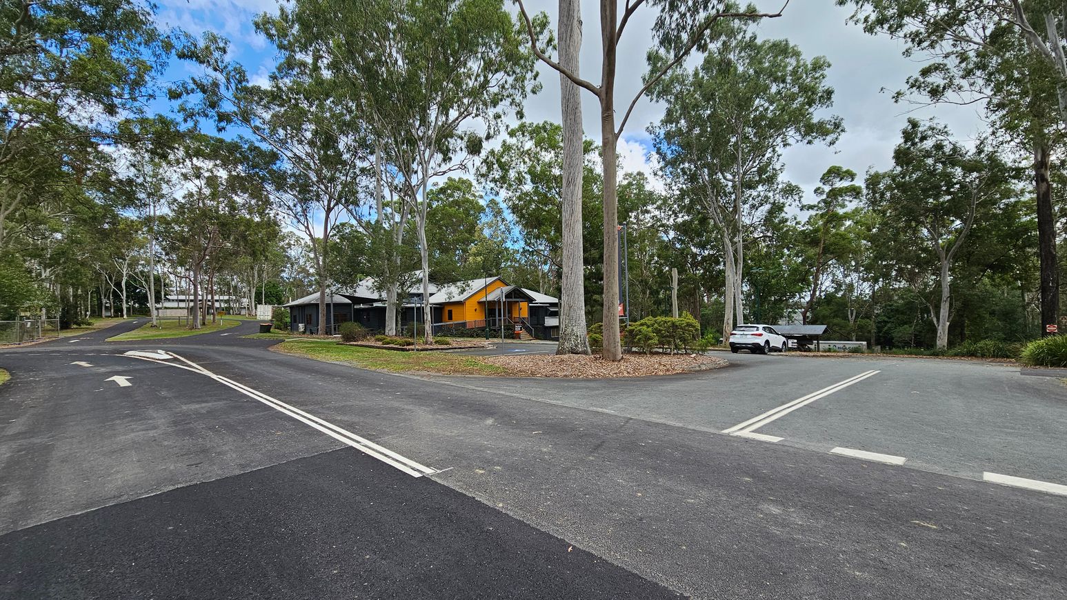 A road forks into two paths surrounded by tall trees. On the left, the road curves uphill with a fence and more trees in the background. In the centre, a building with a bright yellow section is partially visible, surrounded by trees and greenery. On the right, a white car is parked near a small structure, with more trees in the background. The road surface is dark asphalt with white markings.