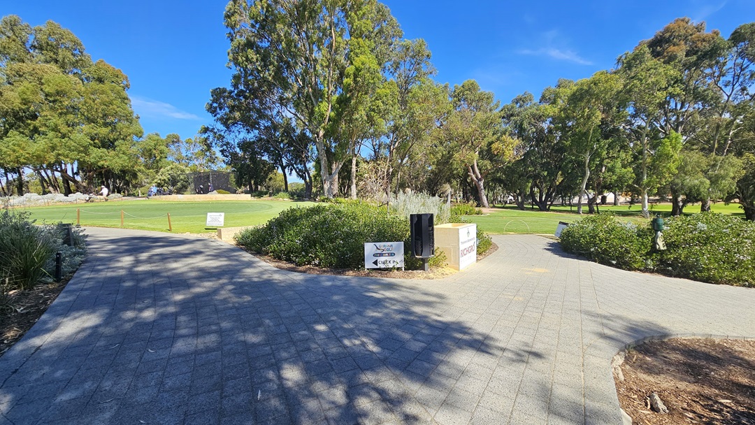 The concrete brick pathway divides; both lead to a vast green and grassy area with tall trees and shrubs. In the middle is a mini landscape with directional signages, a golf hole sponsor, and a garden lamp. The path on the left elevates towards the entrance of the building. A wooden post stands on the right along with a banner. The path on the right descends to the golf area. The concrete brick pathway divides; both lead to a vast green and grassy area with tall trees and shrubs. In the middle is a mini landscape with directional signages, a golf hole sponsor, and a garden lamp. The path on the left elevates towards the entrance of the building. A wooden post stands on the right along with a banner. The path on the right descends to the golf area.