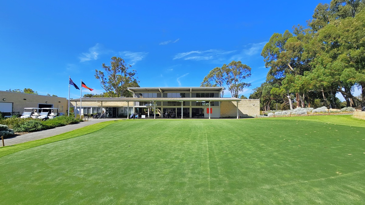 On the right, there is an expansive green space featuring well-maintained grass and synthetic turf. To the left, an ascending path leads to the building. In the middle stands a two-story cream building. Two flags are positioned on the right, accompanied by three golf carts. Shrubs and tall native trees encircle the area. On the right, there is an expansive green space featuring well-maintained grass and synthetic turf. To the left, an ascending path leads to the building. In the middle stands a two-story cream building. Two flags are positioned on the right, accompanied by three golf carts. Shrubs and tall native trees encircle the area.