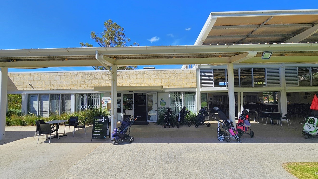The two-level cream building is a modern structure made of brick and glass. In front, there is a shaded facility housing movable plastic tables and chairs, along with a black freestanding banner. Golf carts are visible. The glass entrance is on the left, featuring the facility banner and stickers. More tables and chairs are arranged inside the building on the right. The two-level cream building is a modern structure made of brick and glass. In front, there is a shaded facility housing movable plastic tables and chairs, along with a black freestanding banner. Golf carts are visible. The glass entrance is on the left, featuring the facility banner and stickers. More tables and chairs are arranged inside the building on the right.