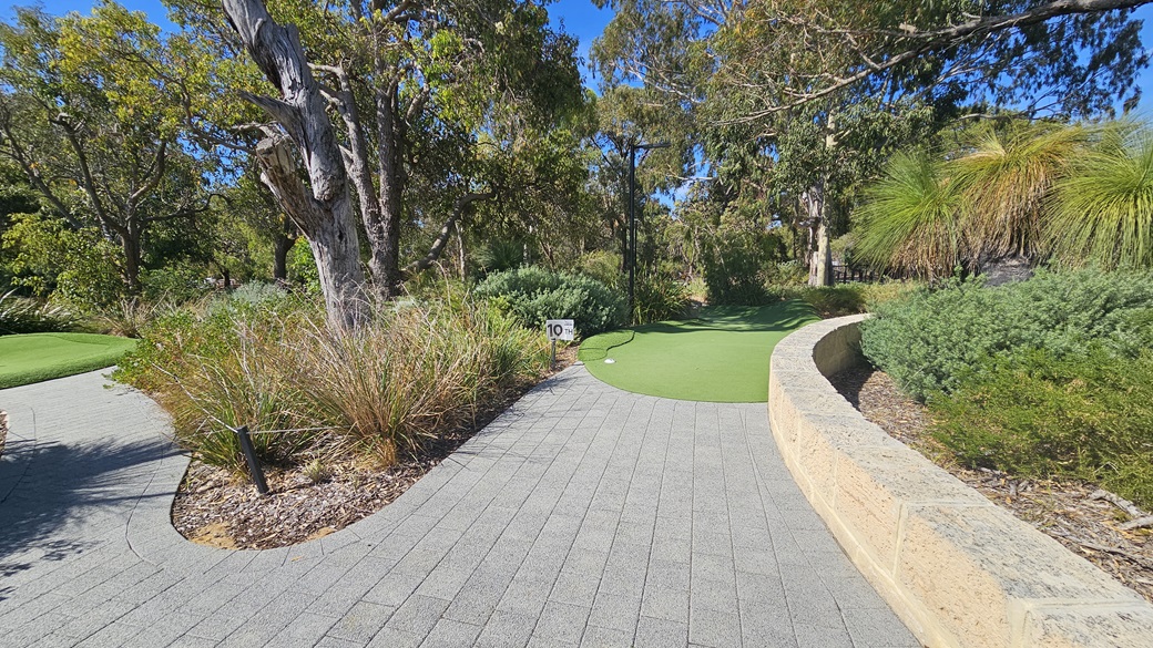 A concrete brick path leading to lower ground. A large, colourful concrete sign is positioned on the right. On the left, a circular brick wall encloses a lamppost. Various types of mature native grass and trees, including Leucophyta shrubs, surround the area. A concrete brick path leading to lower ground. A large, colourful concrete sign is positioned on the right. On the left, a circular brick wall encloses a lamppost. Various types of mature native grass and trees, including Leucophyta shrubs, surround the area.
