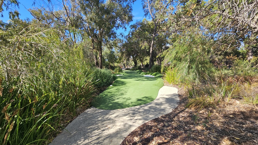 The brick path descends towards the green, surrounded by a landscape of trees, native grass, and shrubs on both sides. To the right, there is a slightly elevated area with soil and native grass. The brick path descends towards the green, surrounded by a landscape of trees, native grass, and shrubs on both sides. To the right, there is a slightly elevated area with soil and native grass.