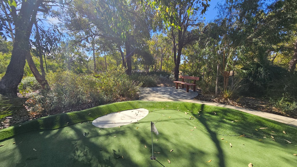 The green is composed of synthetic turf, featuring a bunker in the middle beside a flag. Large trees, native grass, and shrubs encircle the area. A bench is situated beneath a tree in the center. To the right, a descending concrete path is present. The green is composed of synthetic turf, featuring a bunker in the middle beside a flag. Large trees, native grass, and shrubs encircle the area. A bench is situated beneath a tree in the center. To the right, a descending concrete path is present.