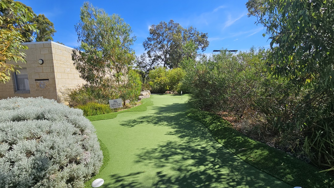 The uneven terrain is paved with concrete bricks and well-kept grass. Ornamental and native plants, along with trees, form a barrier between the grass and concrete. A wooden string fence encloses the area, and a sign reading "1st" is positioned at the center. Tall trees encompass the surroundings. The uneven terrain is paved with concrete bricks and well-kept grass. Ornamental and native plants, along with trees, form a barrier between the grass and concrete. A wooden string fence encloses the area, and a sign reading "1st" is positioned at the center. Tall trees encompass the surroundings.