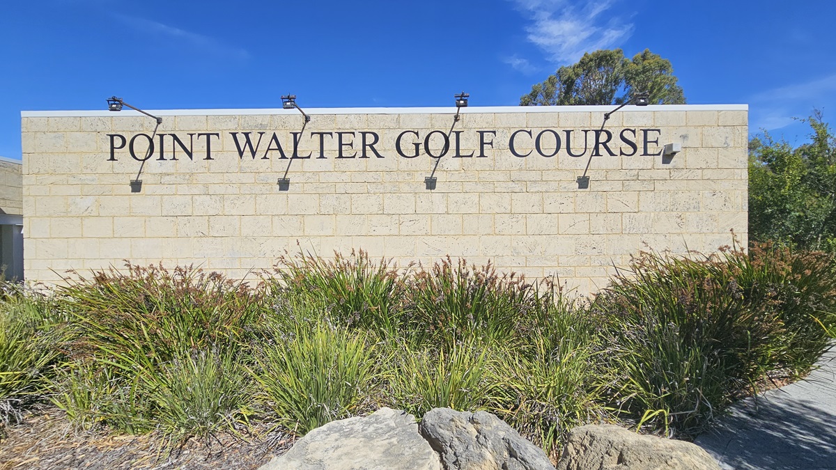 A cream brick building displays a prominent sign reading "Point Walter Golf Course" with four attached lights. Below, there's a landscape of native grass and large rocks. On the right, a brick pathway is situated. A cream brick building displays a prominent sign reading "Point Walter Golf Course" with four attached lights. Below, there's a landscape of native grass and large rocks. On the right, a brick pathway is situated.