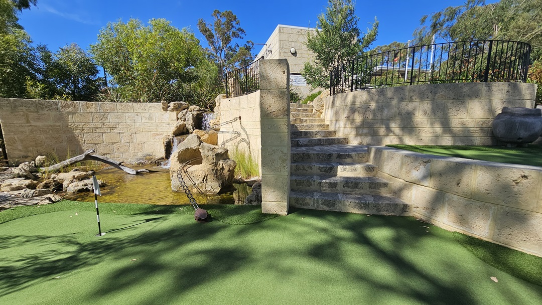 A small waterfall landscape, featuring logs and large rocks, is nestled along a brick wall. To the right are brick stairs leading to a building, surrounded by a metal fence. Well-manicured grass and greenery for the golf hole extend on both the left and right. A banner is positioned on the left, and tall trees shade the area. A small waterfall landscape, featuring logs and large rocks, is nestled along a brick wall. To the right are brick stairs leading to a building, surrounded by a metal fence. Well-manicured grass and greenery for the golf hole extend on both the left and right. A banner is positioned on the left, and tall trees shade the area.