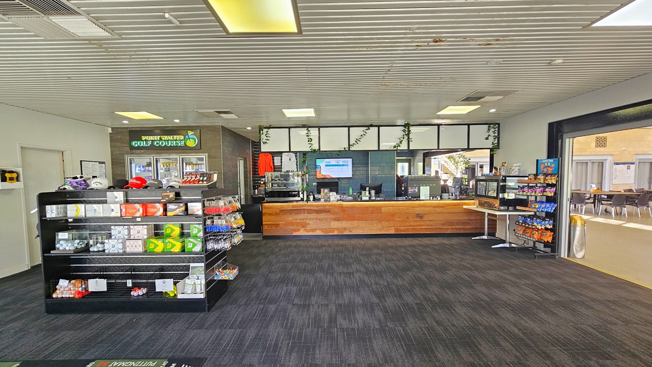 The entrance to the building is through a manual glass door that opens outward. Large banners are affixed to the walls on both sides. A sunscreen dispenser is positioned on the left along with a banner. An anti-slip mat is placed on the doorstep. Native plants are visible on both sides. The ceiling is white with circular fluorescent lights. Inside the facility, the reception area is visible. The entrance to the building is through a manual glass door that opens outward. Large banners are affixed to the walls on both sides. A sunscreen dispenser is positioned on the left along with a banner. An anti-slip mat is placed on the doorstep. Native plants are visible on both sides. The ceiling is white with circular fluorescent lights. Inside the facility, the reception area is visible.