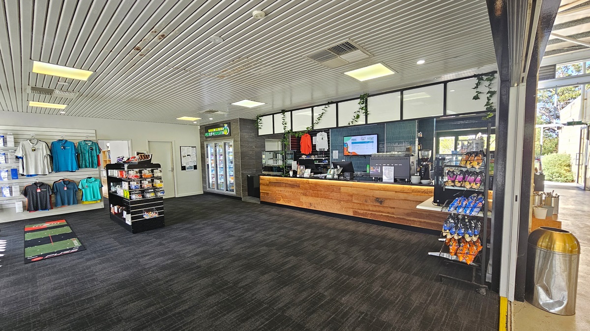 The left side features a display rack with golf items and a large floor mat. The reception desk, with a timber design, showcases retail items and a flat-screen TV. On the counter, a coffee maker and a computer are neatly arranged. Adjacent is a fridge with cold drinks. To the right, a table holds a food rack and a separate one for light snacks. The grey carpeted floor complements the ceiling's corrugated design. Behind a wall, a rubbish bin and a table with kitchen utensils are visible. The left side features a display rack with golf items and a large floor mat. The reception desk, with a timber design, showcases retail items and a flat-screen TV. On the counter, a coffee maker and a computer are neatly arranged. Adjacent is a fridge with cold drinks. To the right, a table holds a food rack and a separate one for light snacks. The grey carpeted floor complements the ceiling's corrugated design. Behind a wall, a rubbish bin and a table with kitchen utensils are visible.