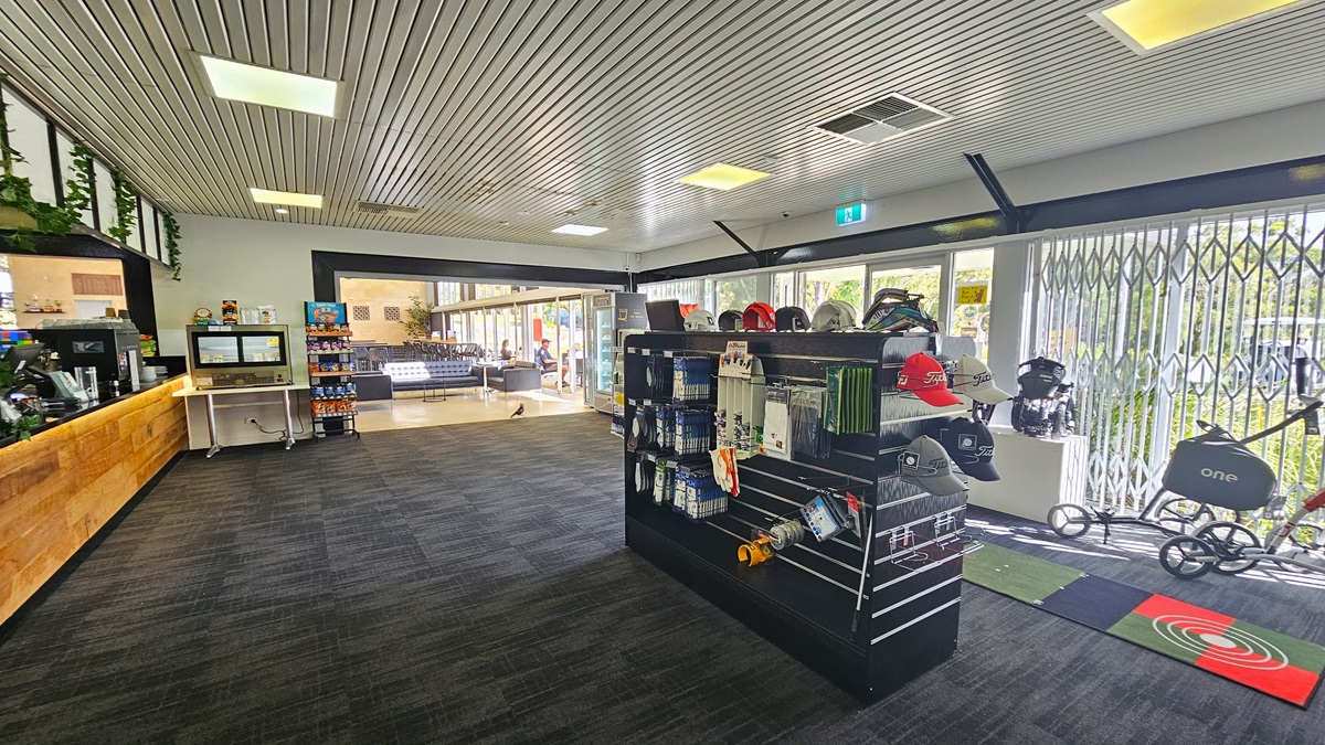 The right side showcases a display rack with golf items and a large floor mat. On the left is the reception desk with a timber design counter. A coffee maker and a computer are neatly arranged on the counter. To the right, a table holds a food rack and a separate one for light snacks. Black leather couches are further in view. The grey carpeted floor complements the ceiling's corrugated design. The right wall is glass with grills. The right side showcases a display rack with golf items and a large floor mat. On the left is the reception desk with a timber design counter. A coffee maker and a computer are neatly arranged on the counter. To the right, a table holds a food rack and a separate one for light snacks. Black leather couches are further in view. The grey carpeted floor complements the ceiling's corrugated design. The right wall is glass with grills.