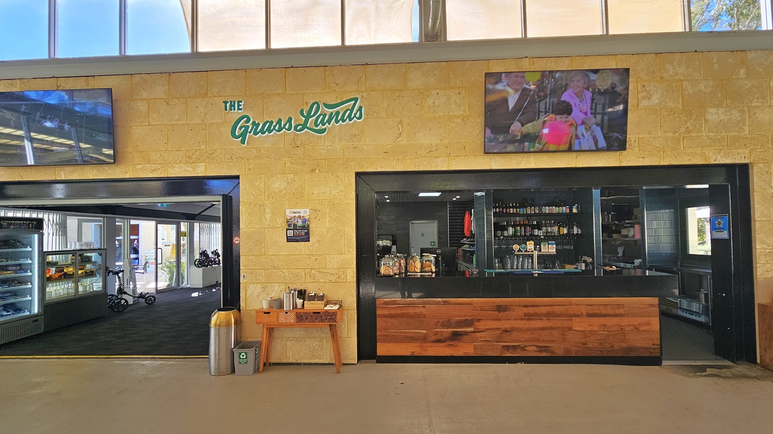 To the left is part of the retail area outside the cafe, while the cafe/bar counter, featuring black walls and a timber-designed countertop, is on the right. Inside the bar, bottles of wine and alcohol are displayed. Two large flat-screen TVs are mounted on the cream wall, showcasing the cafe's brand. A table hosting condiments is placed in the middle, with a silver rubbish bin to its left. To the left is part of the retail area outside the cafe, while the cafe/bar counter, featuring black walls and a timber-designed countertop, is on the right. Inside the bar, bottles of wine and alcohol are displayed. Two large flat-screen TVs are mounted on the cream wall, showcasing the cafe's brand. A table hosting condiments is placed in the middle, with a silver rubbish bin to its left.