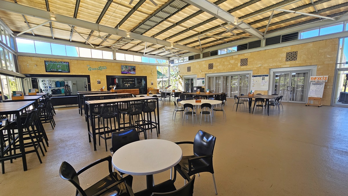 To the left, rows of movable plastic tables and chairs are arranged. On the far left, there's a centre displaying t-shirts for sale. To its right is the cafe and bar counter. Two large flat-screen TVs are mounted on the cream wall. To the right, there are two sets of glass doors. The floor is polished timber, and the ceiling has a sloping industrial design with exposed beams, fluorescent lights, and large ceiling fans. To the left, rows of movable plastic tables and chairs are arranged. On the far left, there's a centre displaying t-shirts for sale. To its right is the cafe and bar counter. Two large flat-screen TVs are mounted on the cream wall. To the right, there are two sets of glass doors. The floor is polished timber, and the ceiling has a sloping industrial design with exposed beams, fluorescent lights, and large ceiling fans.