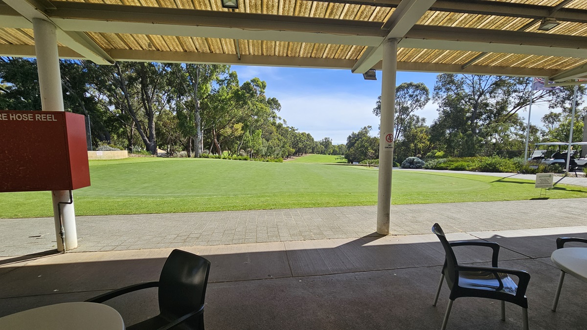 An open space with well-kept green grass. Under the veranda, movable plastic tables and chairs are neatly arranged. The ceiling displays corrugated roofing with exposed metal beams. On the left, a red box houses the fire hose reel. Large trees are visible beyond the grass area. An open space with well-kept green grass. Under the veranda, movable plastic tables and chairs are neatly arranged. The ceiling displays corrugated roofing with exposed metal beams. On the left, a red box houses the fire hose reel. Large trees are visible beyond the grass area.