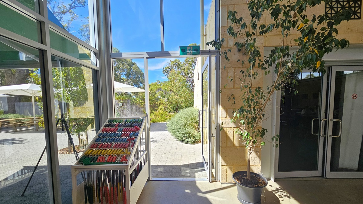 Directly ahead is a glass door which is open and leading to the outside. To the left of this exit are rows of golf clubs lined up by colour and hanging from a metal structure. To the right are glass doors leading to another room. Outside, trees, gardens and umbrellas are visible through the glass windows. Directly ahead is a glass door which is open and leading to the outside. To the left of this exit are rows of golf clubs lined up by colour and hanging from a metal structure. To the right are glass doors leading to another room. Outside, trees, gardens and umbrellas are visible through the glass windows.
