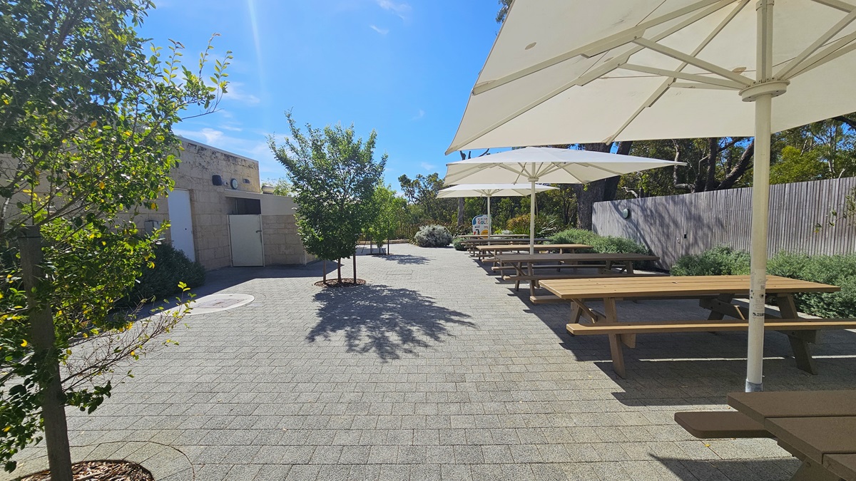 To the left is a low cream building with brick walls. Small trees line the brick pathway. On the right, there's a shaded patio with white umbrella shades and a row of picnic tables. The wall on the right is bordered by plants. To the left is a low cream building with brick walls. Small trees line the brick pathway. On the right, there's a shaded patio with white umbrella shades and a row of picnic tables. The wall on the right is bordered by plants.