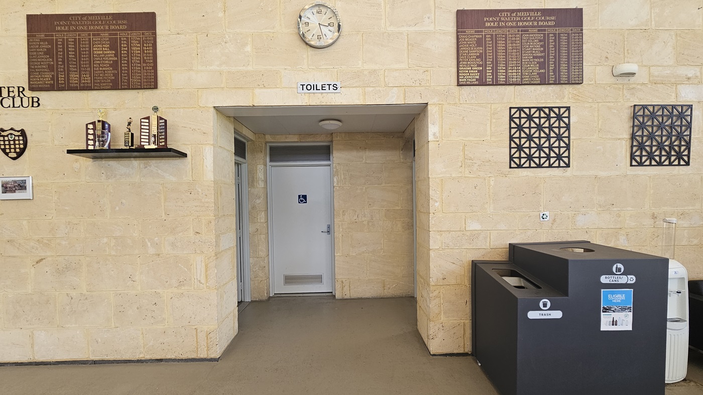 The accessible bathroom, marked with a white door and signage. To the right is a black rubbish bin with a separate hole for recycling. The entire wall is made of cream bricks. Trophies, awards, a brown board featuring "Hole In One Honour Board," a round clock, and wall decorations are displayed. The accessible bathroom, marked with a white door and signage. To the right is a black rubbish bin with a separate hole for recycling. The entire wall is made of cream bricks. Trophies, awards, a brown board featuring "Hole In One Honour Board," a round clock, and wall decorations are displayed.