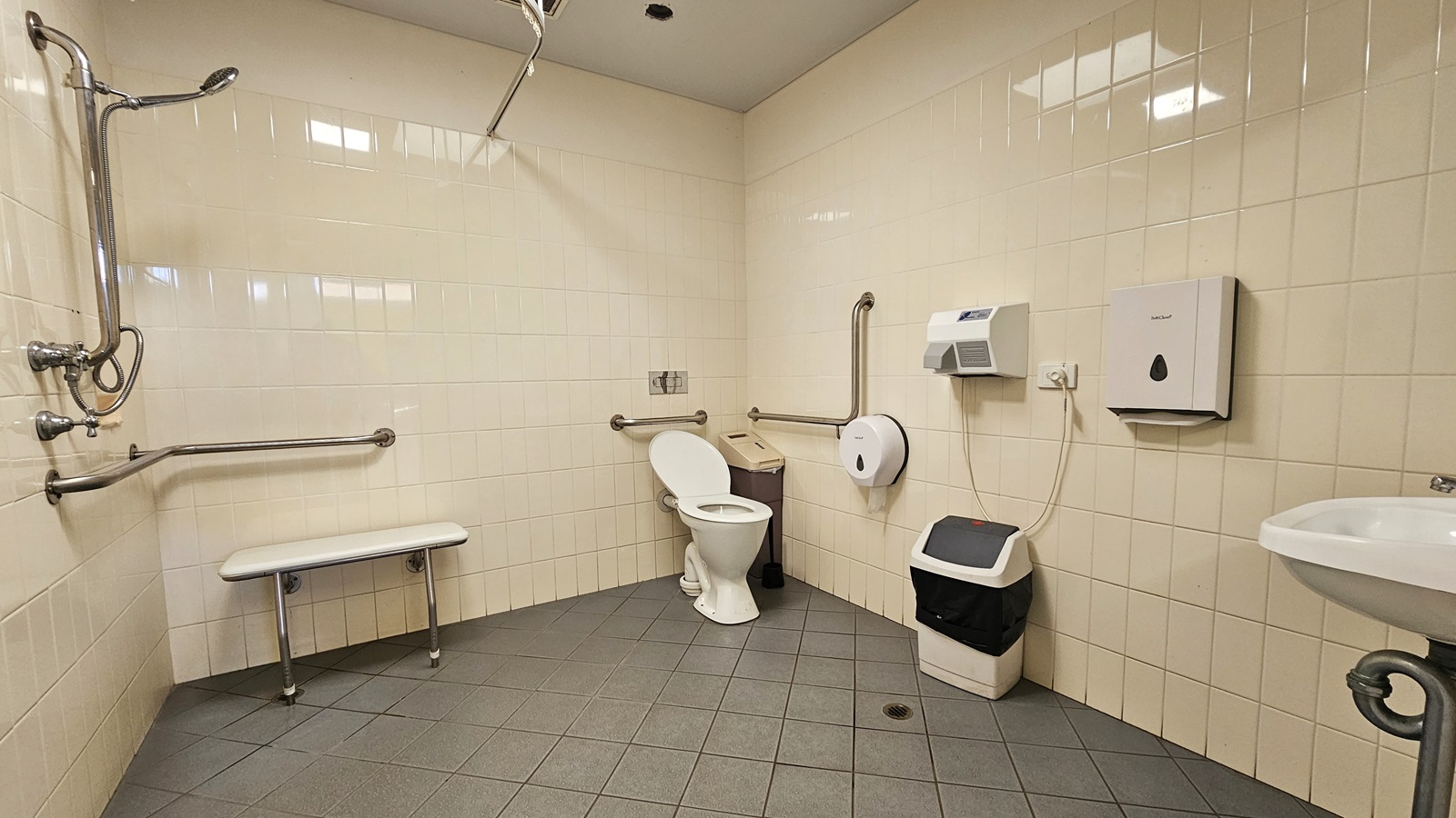 The accessible bathroom restroom features a left-installed shower with accompanying handrails. Adjacent to it, a bench chair is placed. In the corner, a toilet bowl and rubbish bin are situated. On the wall, a tissue dispenser, two rubbish bins, and a hand dryer are affixed, alongside a wash basin. The wall exhibits white tiles, and the ceiling and floor are adorned with white and grey tiles, respectively. The accessible bathroom restroom features a left-installed shower with accompanying handrails. Adjacent to it, a bench chair is placed. In the corner, a toilet bowl and rubbish bin are situated. On the wall, a tissue dispenser, two rubbish bins, and a hand dryer are affixed, alongside a wash basin. The wall exhibits white tiles, and the ceiling and floor are adorned with white and grey tiles, respectively.