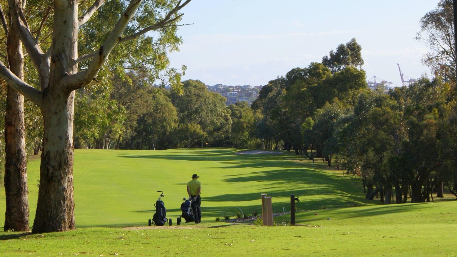 The golf course area offers a scenic view with a hillside and a green consisting of natural grass and synthetic turfs. On the left stands a mature tree. To the right, two concrete bollards flank a garden lamp post. Additional trees are situated on the right. In the middle, a golfer stands with a golf bag nearby. The golf course area offers a scenic view with a hillside and a green consisting of natural grass and synthetic turfs. On the left stands a mature tree. To the right, two concrete bollards flank a garden lamp post. Additional trees are situated on the right. In the middle, a golfer stands with a golf bag nearby.