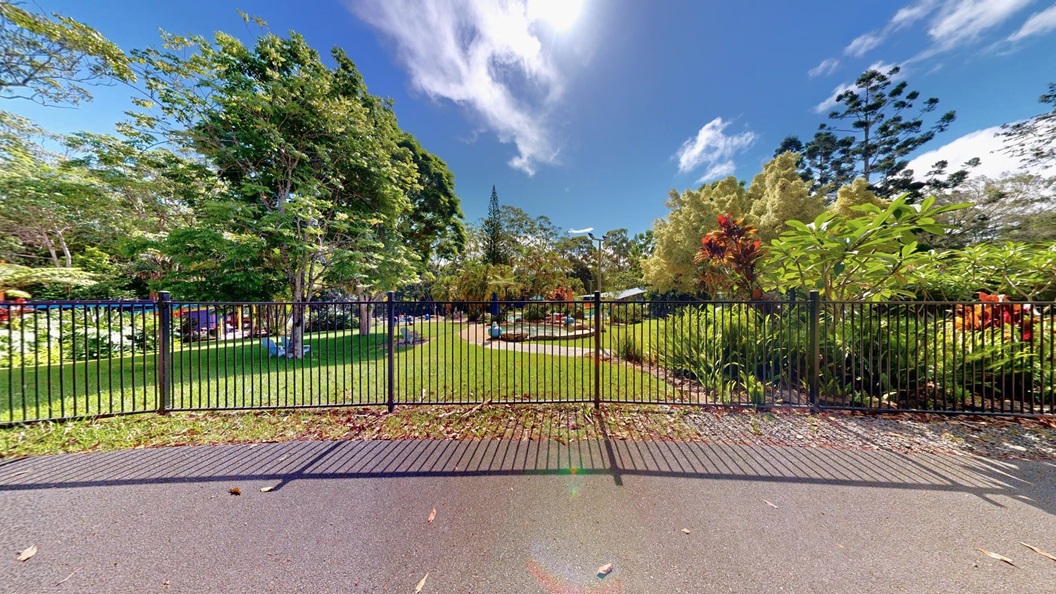 A black metal fence divides the aquatic area from the road. Inside, pools are visible. Plastic chairs are under tree shades on the left, while various trees, ferns, and ornamental plants decorate the landscape on the right. A black metal fence divides the aquatic area from the road. Inside, pools are visible. Plastic chairs are under tree shades on the left, while various trees, ferns, and ornamental plants decorate the landscape on the right.