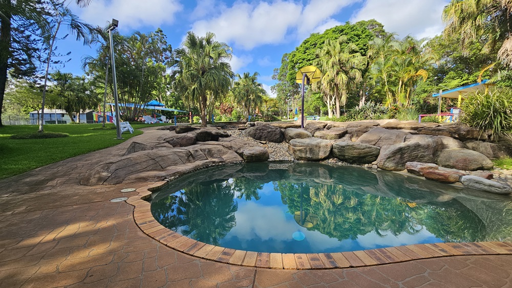 A circular pool is bordered by large rocks and pebbles. Plastic lounge chairs are placed on the grass area to the left, alongside palm trees. Trees and grass encircle the area. A basketball board is on the pool deck. On the right, there's a colourful shade structure. Trees surround the area. A circular pool is bordered by large rocks and pebbles. Plastic lounge chairs are placed on the grass area to the left, alongside palm trees. Trees and grass encircle the area. A basketball board is on the pool deck. On the right, there's a colourful shade structure. Trees surround the area.