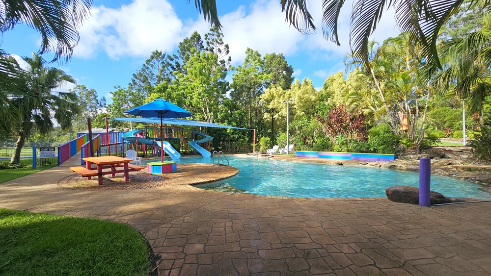 A children's pool with handrails and steps. Blue slides with multicolored ramps on the left, featuring safety reminders. A picnic table and umbrella shade on the pool deck. A large rock and a purple bollard are visible. Surrounding the area are various trees and grass. Plastic lounge chairs are positioned on the other side of the pool. A children's pool with handrails and steps. Blue slides with multicolored ramps on the left, featuring safety reminders. A picnic table and umbrella shade on the pool deck. A large rock and a purple bollard are visible. Surrounding the area are various trees and grass. Plastic lounge chairs are positioned on the other side of the pool.