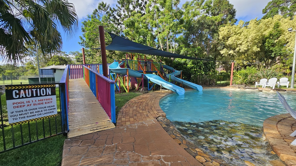 A children's pool with handrails and steps. Blue slides with multicolored ramps on the left, featuring safety reminders. A black shade cloth hangs overhead. Surrounding the area are various trees and grass. Plastic lounge chairs are positioned on the other side of the pool. A children's pool with handrails and steps. Blue slides with multicolored ramps on the left, featuring safety reminders. A black shade cloth hangs overhead. Surrounding the area are various trees and grass. Plastic lounge chairs are positioned on the other side of the pool.