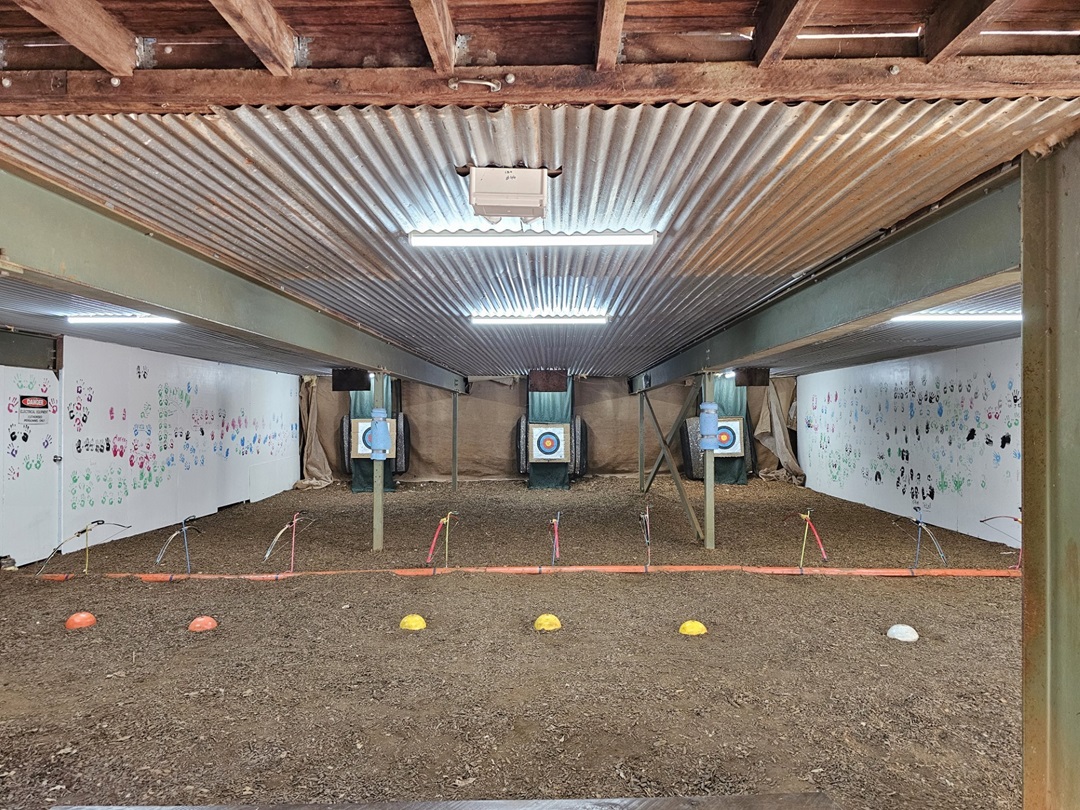 On the left and right, white walls are covered with various colourful handprints and drawings. In the middle, there are three archery targets mounted on stands, each with a blue and white bullseye. The background features a beige netting behind the targets. The foreground has a dirt floor with a row of yellow, white, and orange cones. The ceiling is corrugated metal with exposed wooden beams. On the left and right, white walls are covered with various colourful handprints and drawings. In the middle, there are three archery targets mounted on stands, each with a blue and white bullseye. The background features a beige netting behind the targets. The foreground has a dirt floor with a row of yellow, white, and orange cones. The ceiling is corrugated metal with exposed wooden beams.
