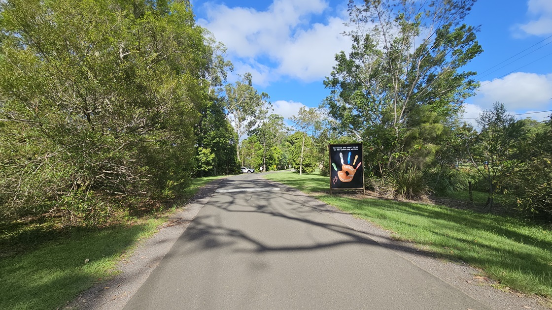 A straight bitumen road. Alongside the road, there is a large sign situated on the grassy area to the right. The road is flanked on both sides by various trees with robust branches and grass. A straight bitumen road. Alongside the road, there is a large sign situated on the grassy area to the right. The road is flanked on both sides by various trees with robust branches and grass.
