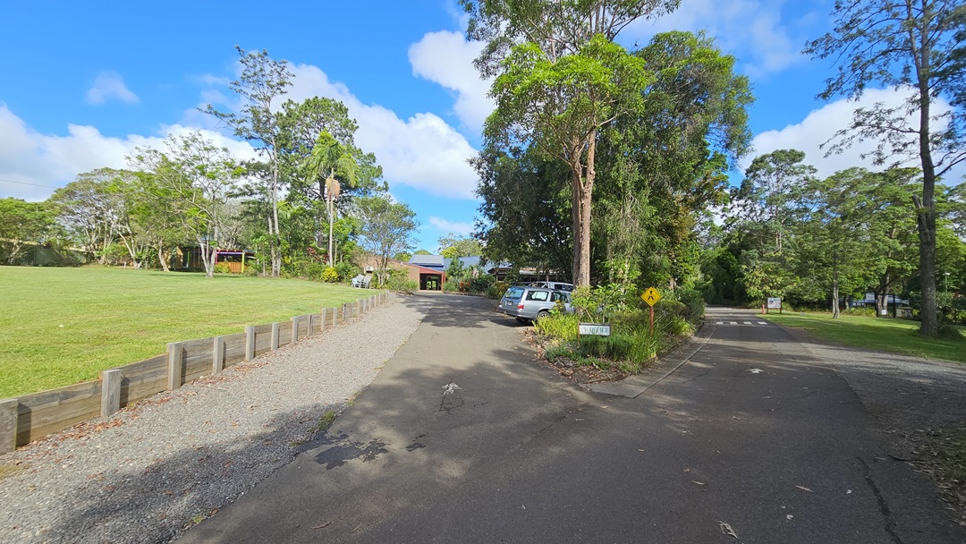 The road splits: To the left, it leads to a car park and a building with a brown brick facade, alongside a field of manicured grass bordered by a low concrete fence. The right-hand road leads to an open area surrounded by trees and grass. In the middle aisle, there is a sign, set against a backdrop of tall trees and grass. The road splits: To the left, it leads to a car park and a building with a brown brick facade, alongside a field of manicured grass bordered by a low concrete fence. The right-hand road leads to an open area surrounded by trees and grass. In the middle aisle, there is a sign, set against a backdrop of tall trees and grass.