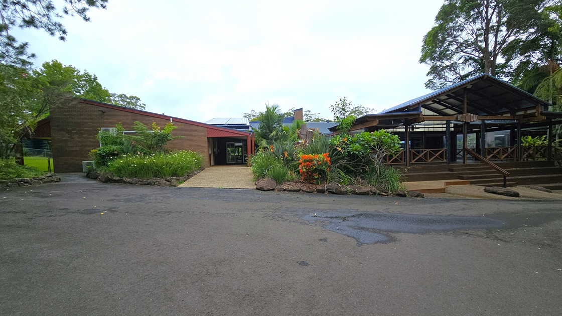 To the left, there's a low-rise building with a brown concrete facade and pitched roofing. It is surrounded by a landscaped garden with colourful flowering plants. On the right is another building with a pitched corrugated roof, accessible via concrete steps with railings. Wooden railings enclose this area. Tall trees of various types surround the vicinity. To the left, there's a low-rise building with a brown concrete facade and pitched roofing. It is surrounded by a landscaped garden with colourful flowering plants. On the right is another building with a pitched corrugated roof, accessible via concrete steps with railings. Wooden railings enclose this area. Tall trees of various types surround the vicinity.