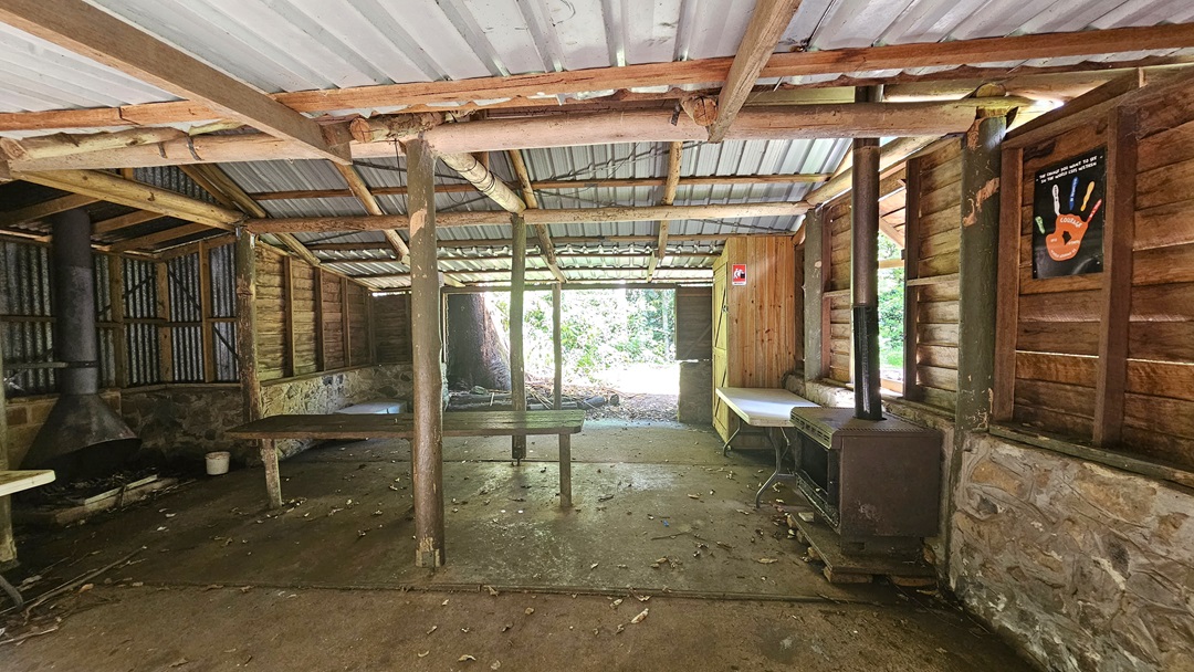 A hut with a half-concrete, half-timber structure. The corrugated roof and wooden beams make up the ceiling. A poster hangs on the right timber wall. Tables, bench chairs, and a cabinet are arranged against the wall. A fireplace and chimney are on the left. The uneven floor is hard concrete with scattered dry foliage. A hut with a half-concrete, half-timber structure. The corrugated roof and wooden beams make up the ceiling. A poster hangs on the right timber wall. Tables, bench chairs, and a cabinet are arranged against the wall. A fireplace and chimney are on the left. The uneven floor is hard concrete with scattered dry foliage.