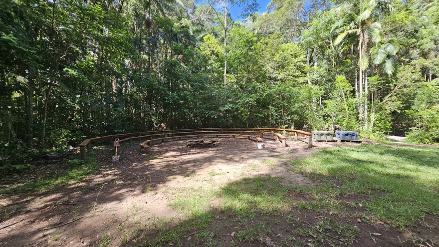 In an open unpaved area partially covered with grass, wooden bench chairs form a U-shape on the left. Tall trees, bushes, native plants, and grass, including palm trees, border the back. In an open unpaved area partially covered with grass, wooden bench chairs form a U-shape on the left. Tall trees, bushes, native plants, and grass, including palm trees, border the back.