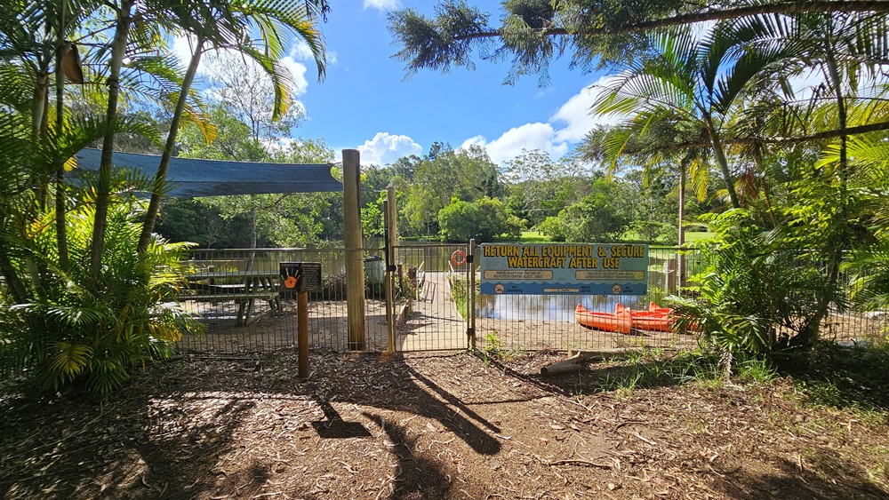 Bench chairs are neatly arranged beneath a shade cloth secured by a metal fence displaying posters and facility reminders. A wooden platform extends from the shade to the lake. Orange watercraft rest by the lake on the right, bordered by palm trees in the front and various trees surrounding the area. The ground is unpaved. Bench chairs are neatly arranged beneath a shade cloth secured by a metal fence displaying posters and facility reminders. A wooden platform extends from the shade to the lake. Orange watercraft rest by the lake on the right, bordered by palm trees in the front and various trees surrounding the area. The ground is unpaved.