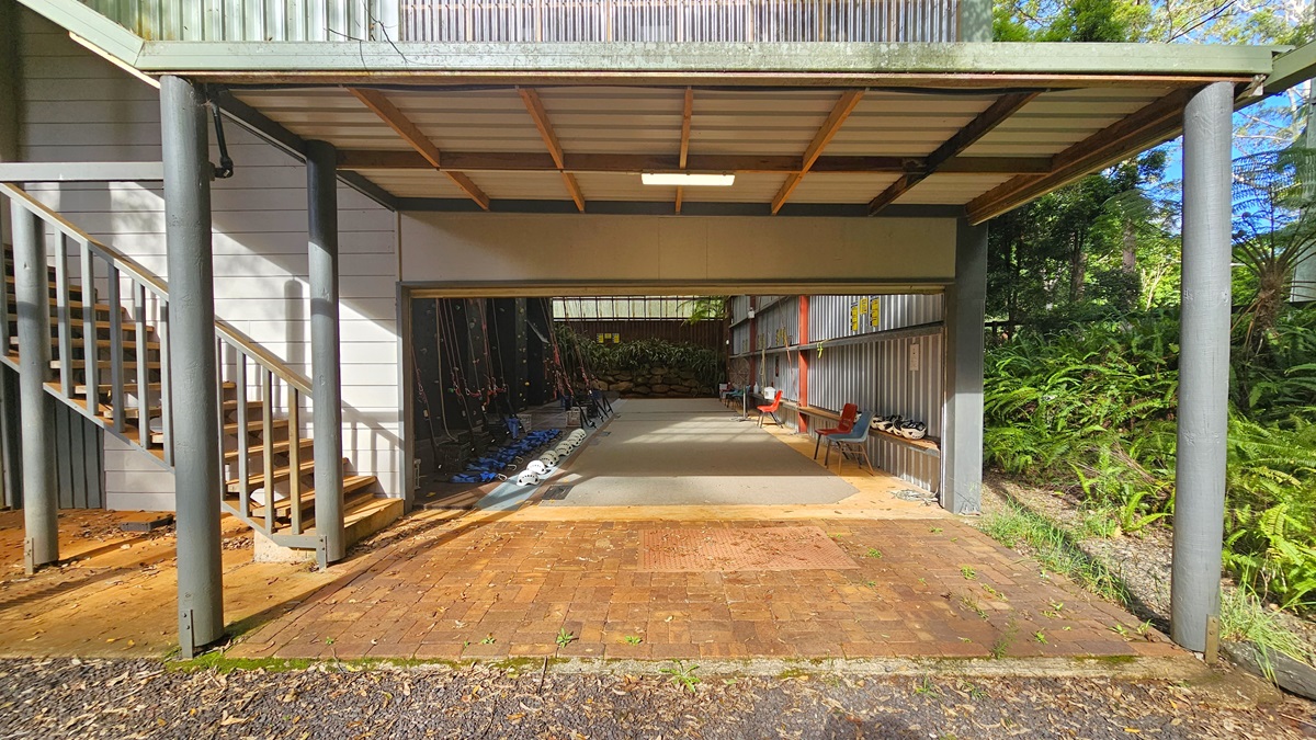 To the left is a staircase with metal handrails. Under the shade of the tower are climbing gear such as ropes and helmets positioned on the floor in front of the black climbing wall. Plastic chairs are lined near the corrugated wall, along with headgear. Landscape plants fill the end. The area on the right is filled with native grass, ferns, and trees. The floor has concrete and bricks. To the left is a staircase with metal handrails. Under the shade of the tower are climbing gear such as ropes and helmets positioned on the floor in front of the black climbing wall. Plastic chairs are lined near the corrugated wall, along with headgear. Landscape plants fill the end. The area on the right is filled with native grass, ferns, and trees. The floor has concrete and bricks.