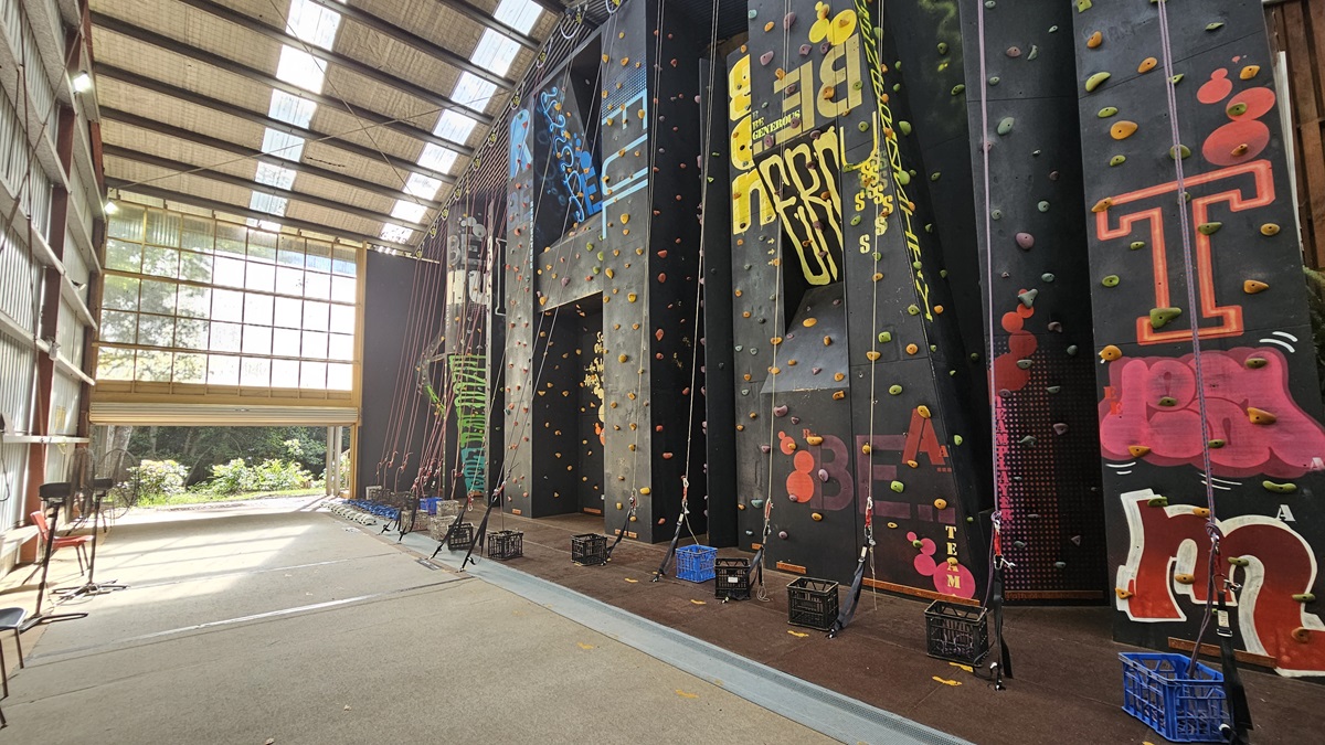 To the right, a black climbing wall with patterns and multi-colored steps is equipped with climbing gear like ropes, headgear, and gloves. The ceiling and upper wall display large metal beams with silver insulation. The left wall has corrugated metal sheets. Plastic chairs are arranged, along with a fan. The industrial door is rolled up at the top. To the right, a black climbing wall with patterns and multi-colored steps is equipped with climbing gear like ropes, headgear, and gloves. The ceiling and upper wall display large metal beams with silver insulation. The left wall has corrugated metal sheets. Plastic chairs are arranged, along with a fan. The industrial door is rolled up at the top.